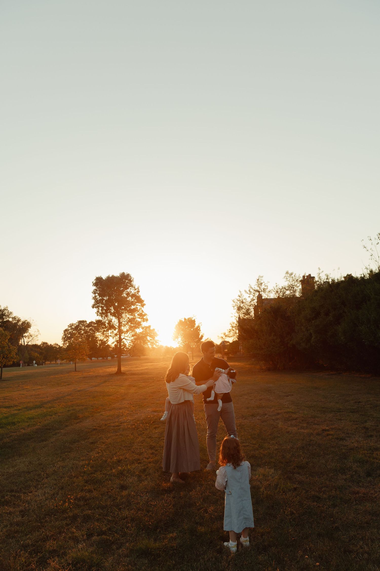 Family silhouetted against golden sunset in park, two adults holding children while toddler stands nearby on grassy field.
