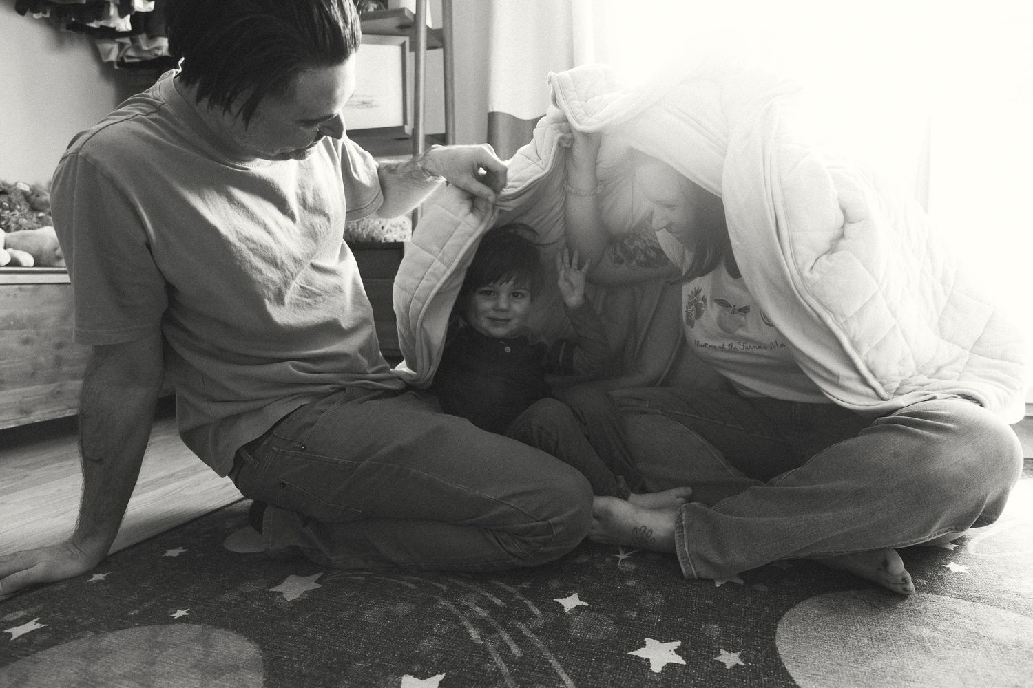 Black and white photo of two parents huddled over a young child sitting on a star-patterned rug indoors.