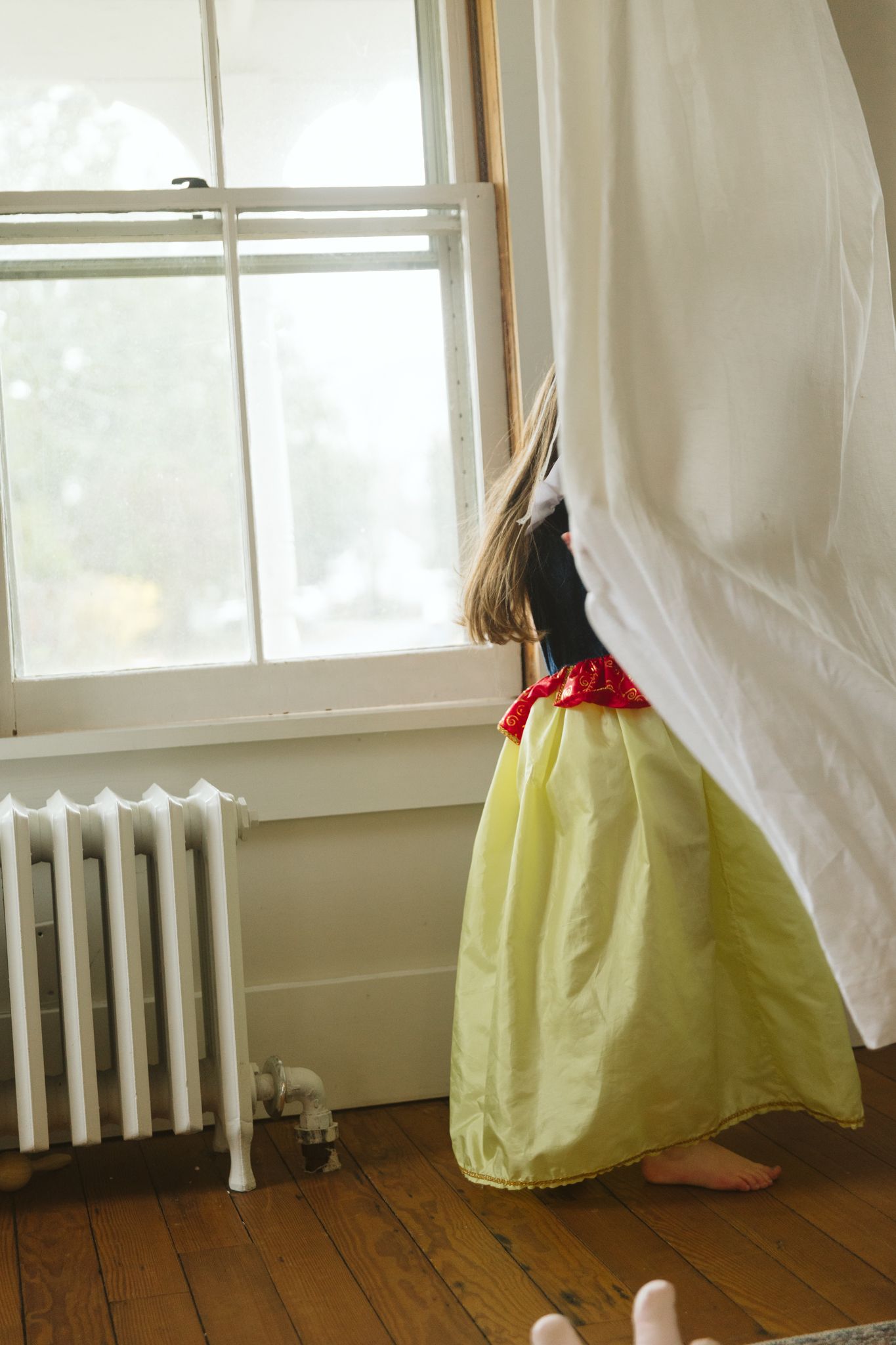 White curtains and yellow skirt shown against wooden floors and radiator near a window.
