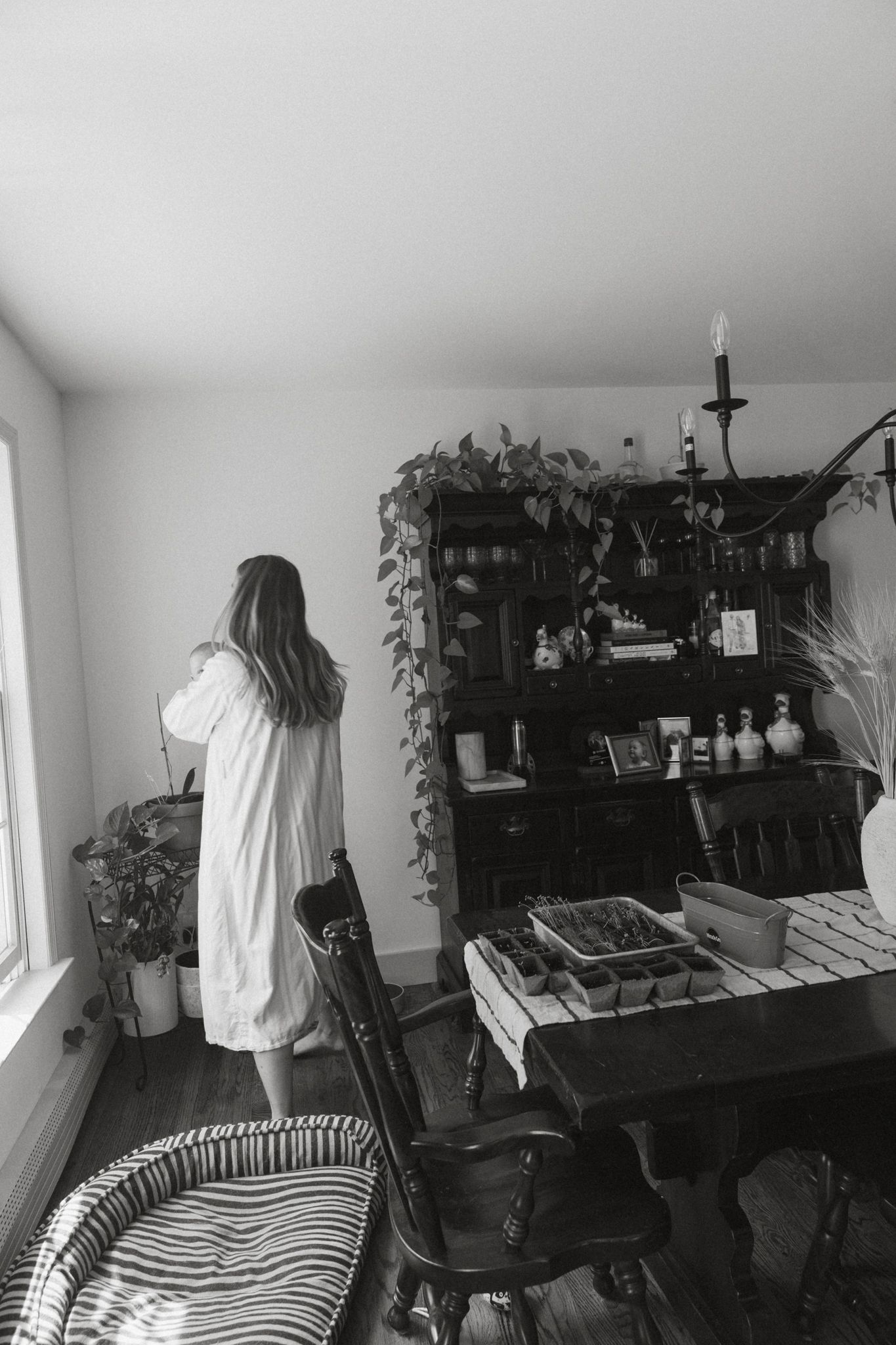 Woman in white robe standing by window in vintage dining room with dark wood hutch and houseplants.