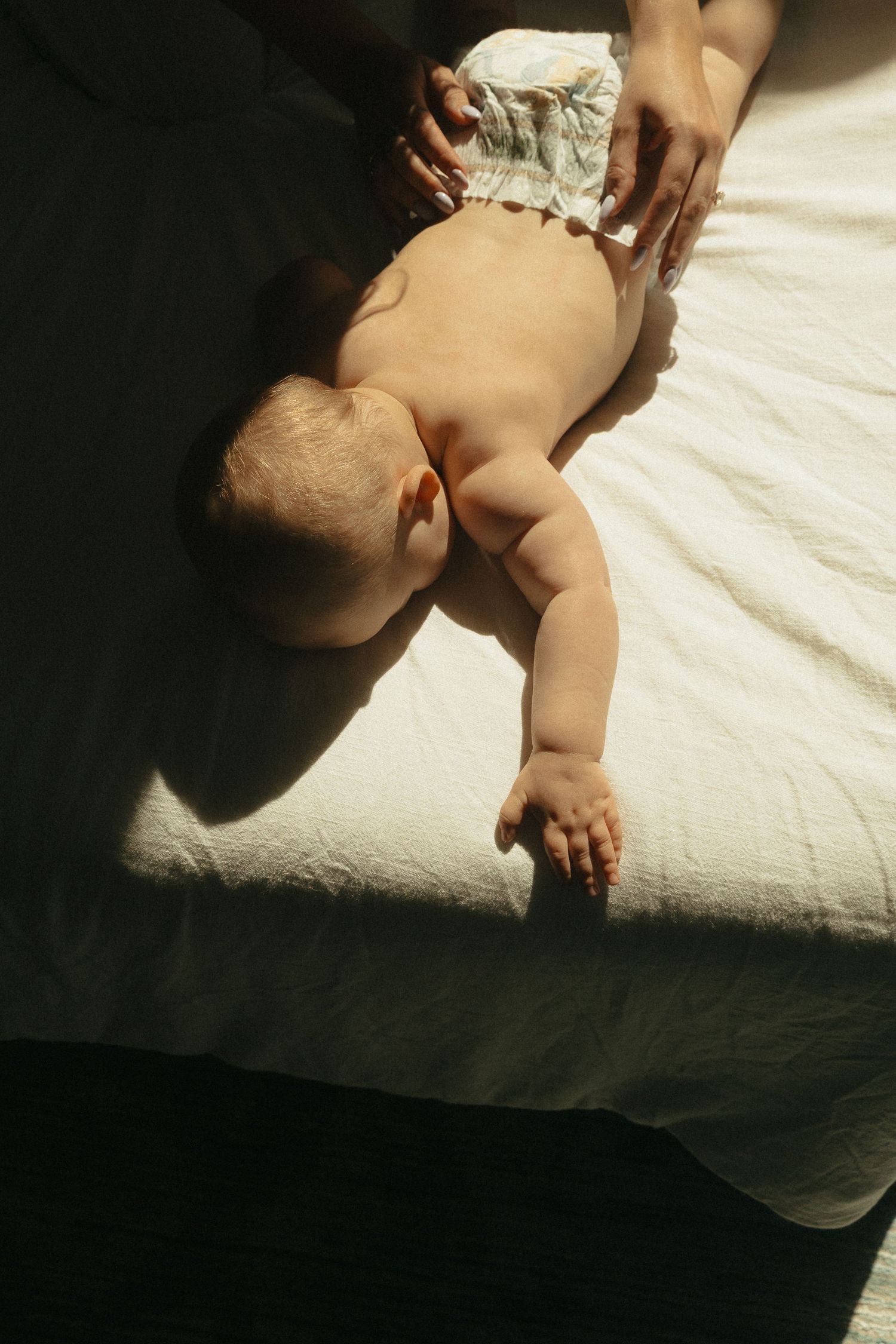 Baby lying on stomach in diaper on white bed, with adult hands gently supporting in soft sunlight.