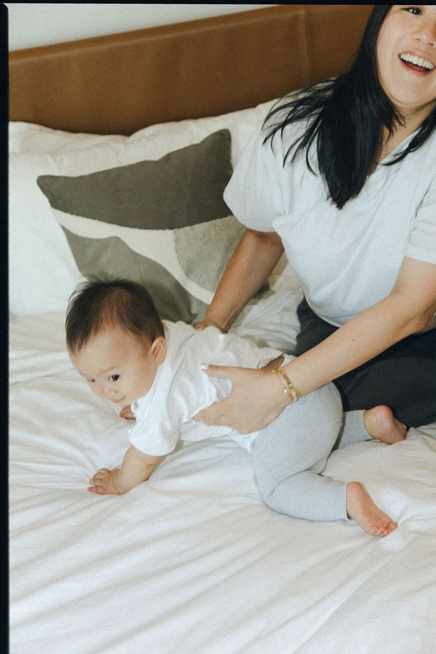 Smiling mother in grey outfit helping baby crawl on white bedding with neutral pillow backdrop.