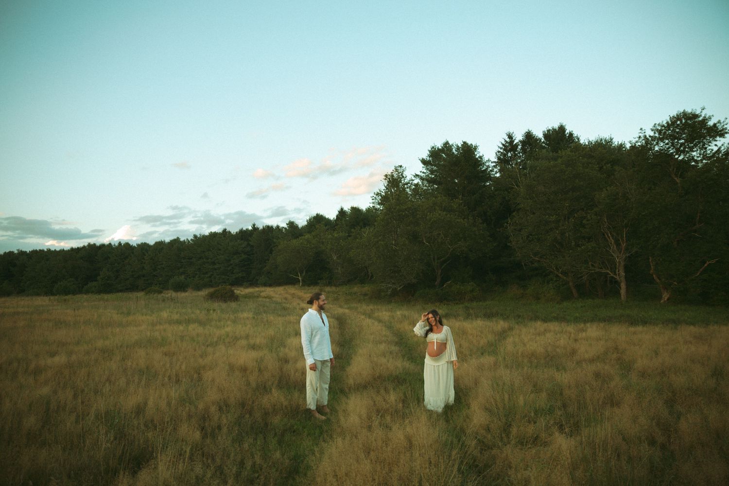 Couple standing apart in golden field at dusk with lush forest backdrop, romantic outdoor portrait photography.