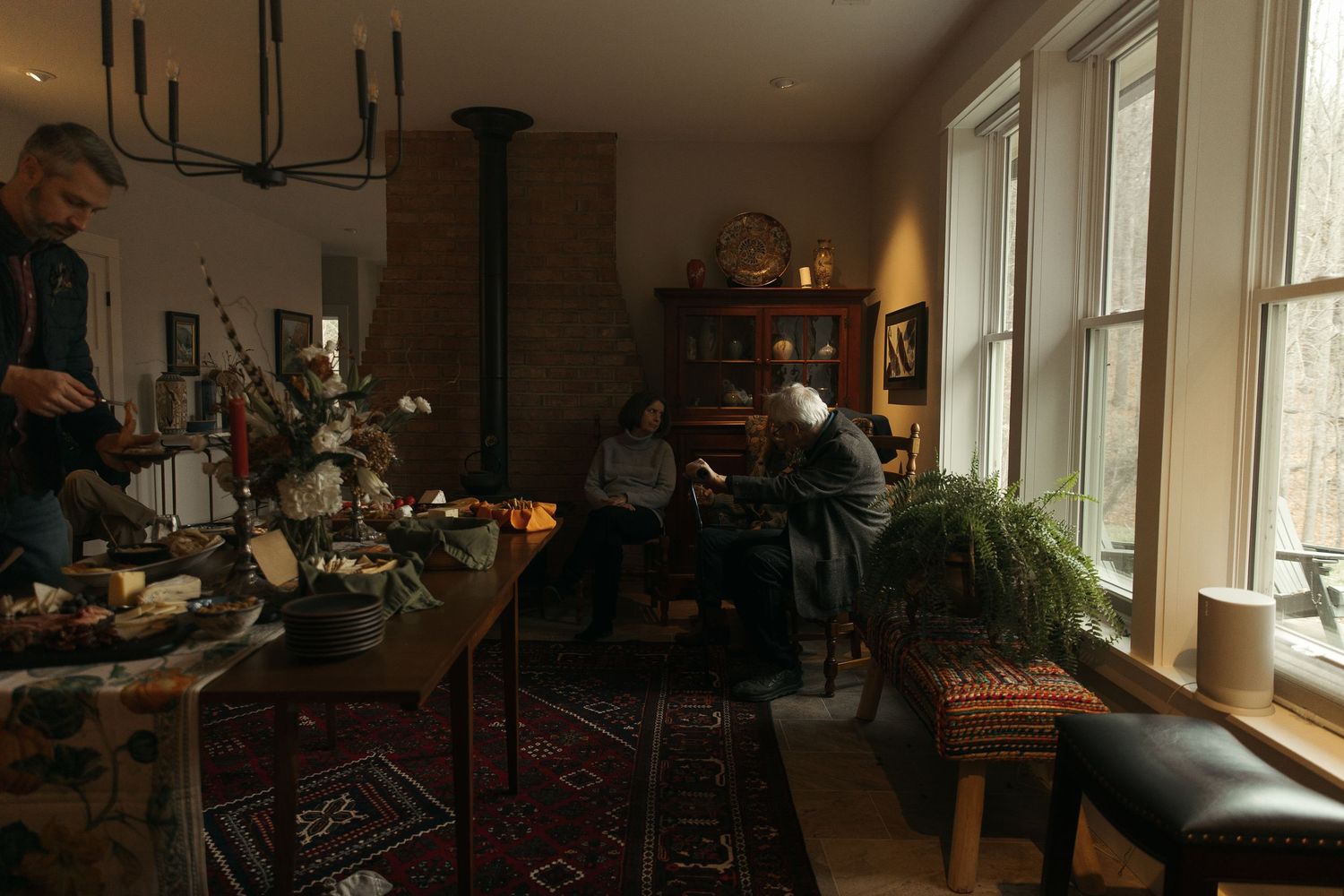 Cozy living room gathering with people relaxing near a wood stove, antique furniture, and large windows with natural light.
