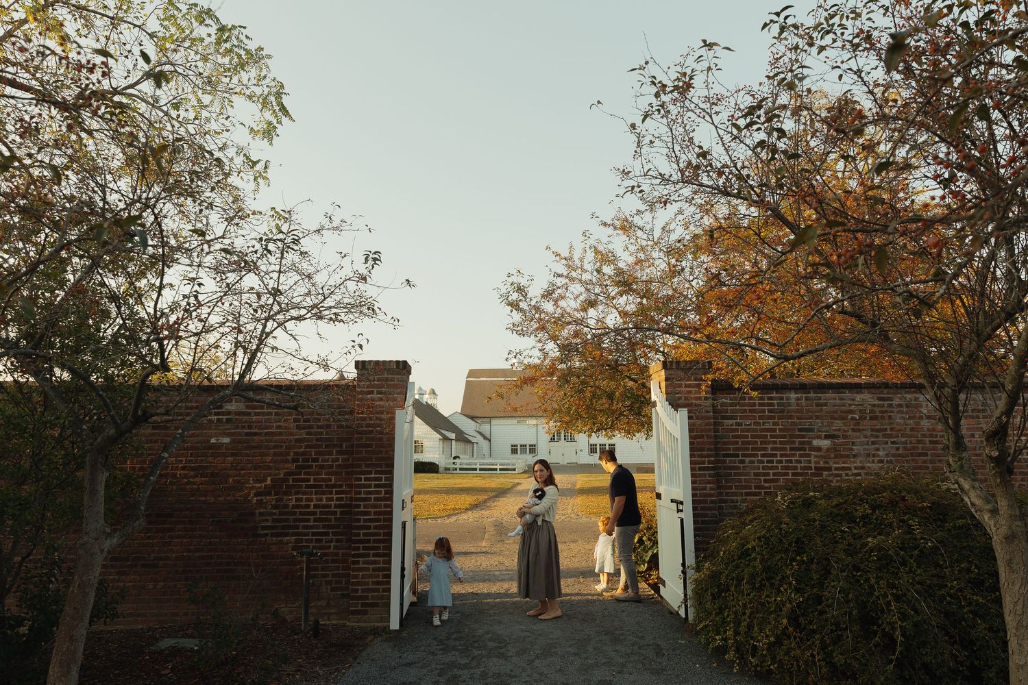 Family walking through white gate entrance of brick wall at golden hour farm.