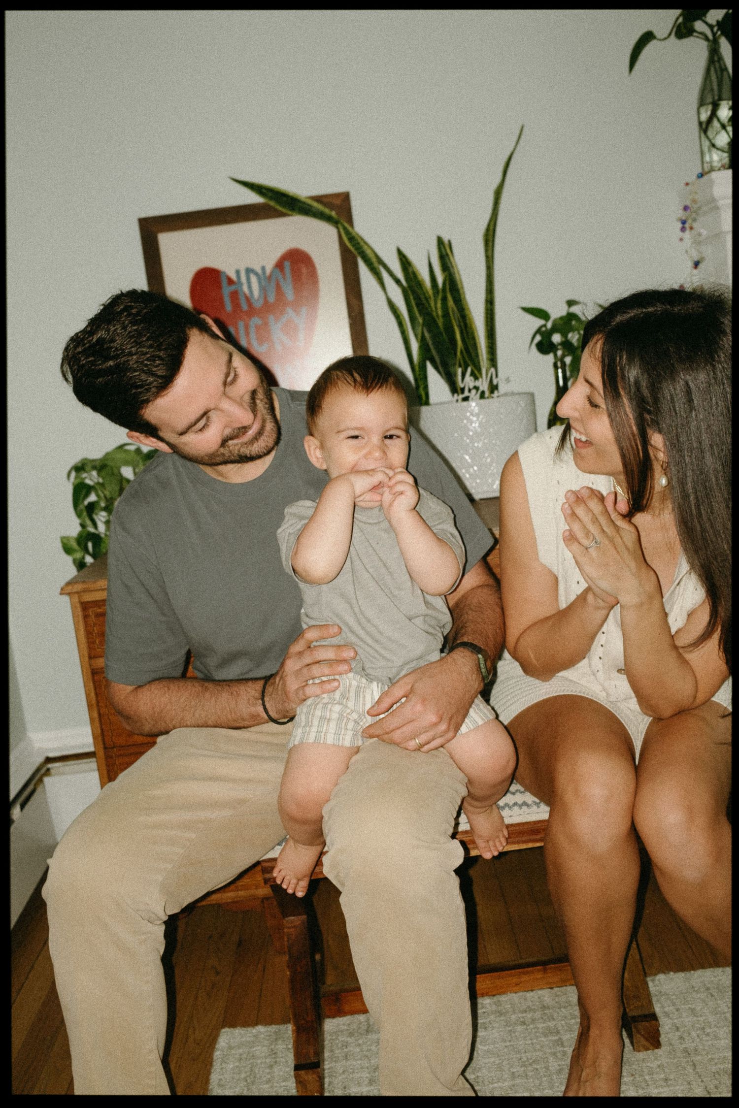 Young family with baby sitting on wooden bench indoors, parents smiling at infant in gray onesie.