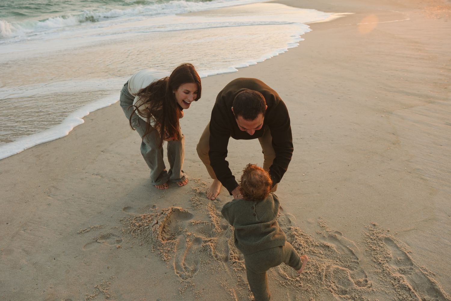 Parents playing with their child on a sandy beach at sunset.