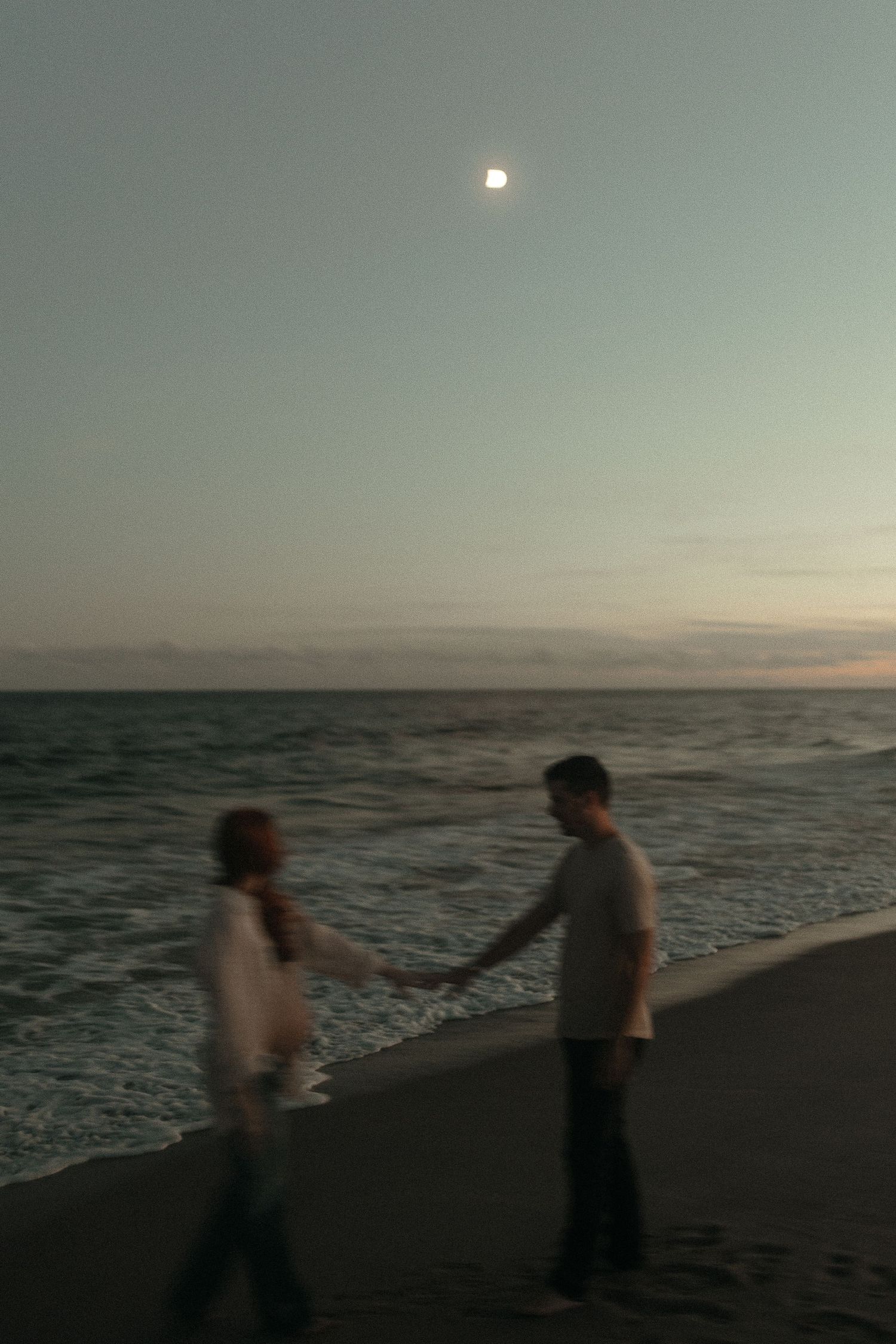 Two silhouettes hold hands on a beach at sunset with a bright moon in the sky.