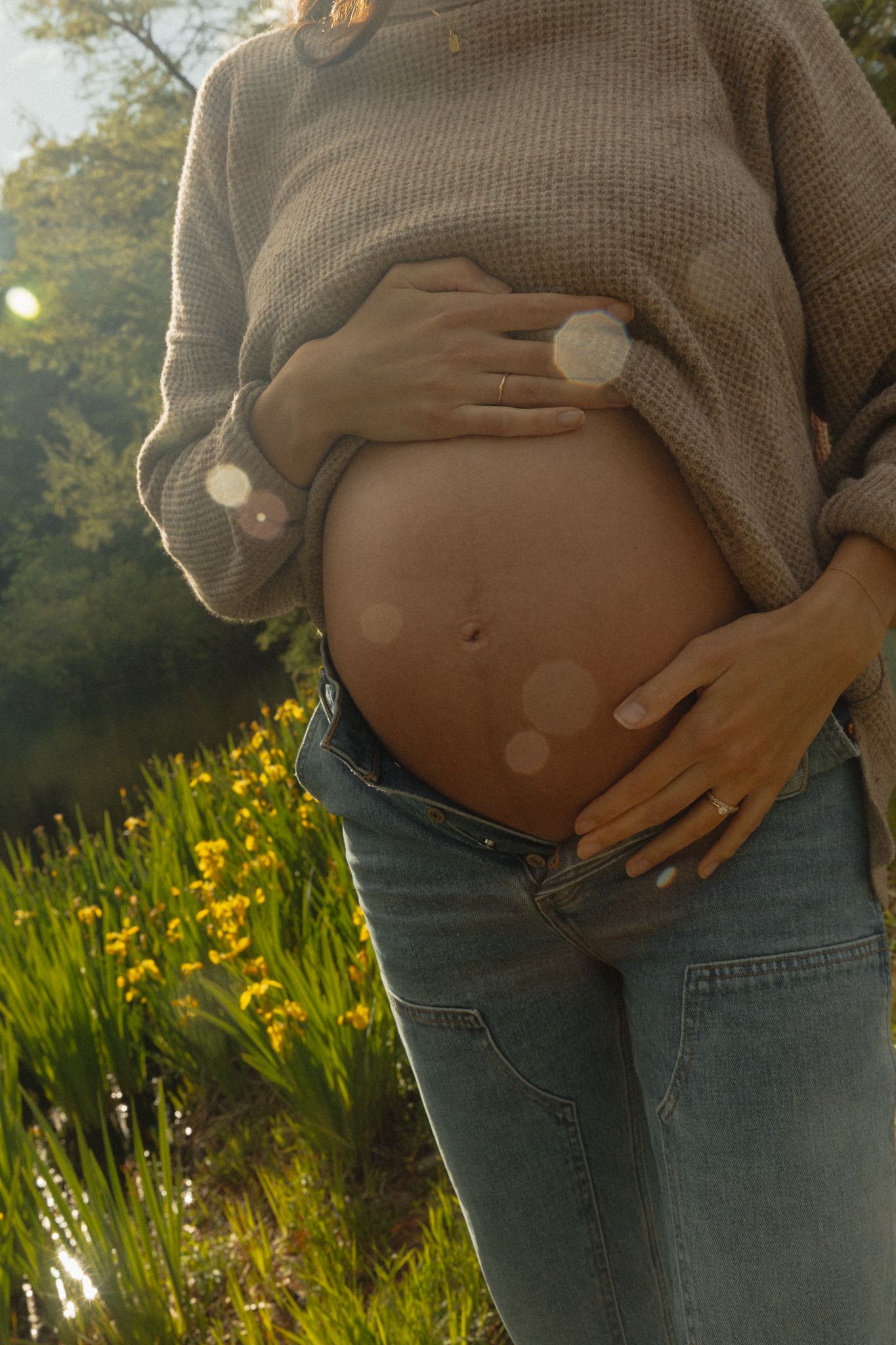 Pregnant woman cradles bare baby bump outdoors near yellow wildflowers in golden sunlight.
