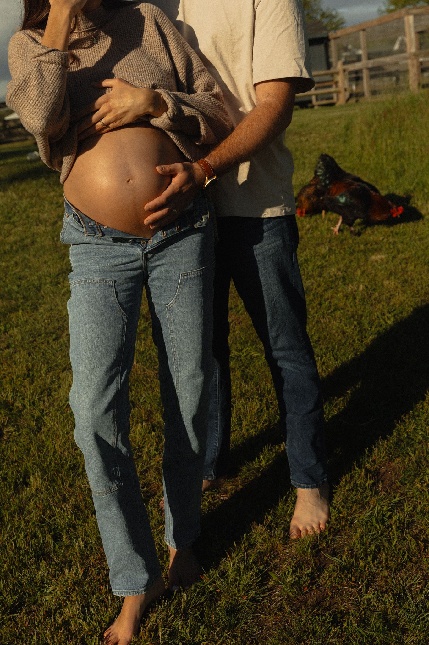Couple on a farm at golden hour with man embracing pregnant woman from behind.