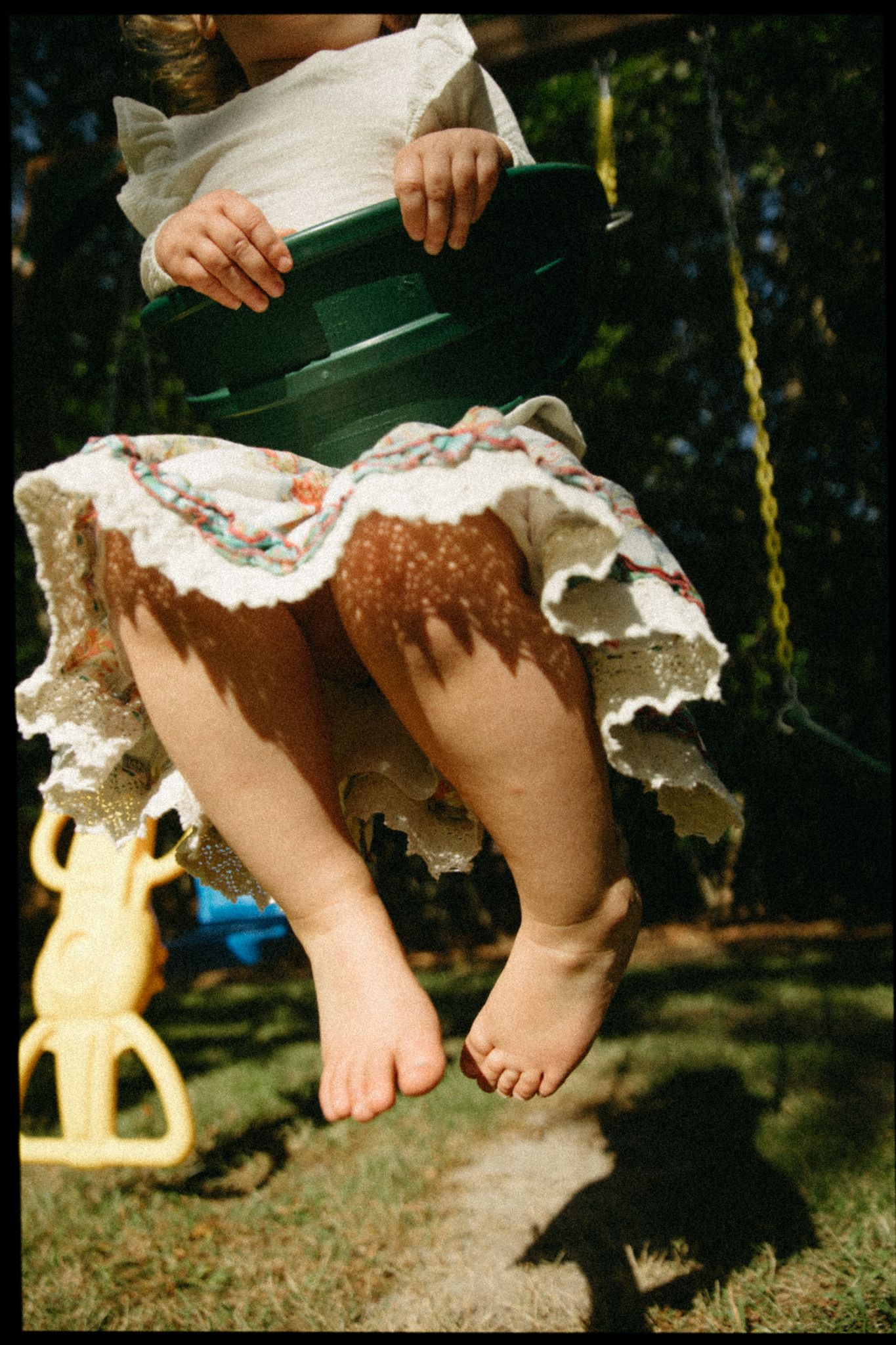 Young child sitting on a green swing outdoors, wearing a white lace dress with bare feet dangling above grass.