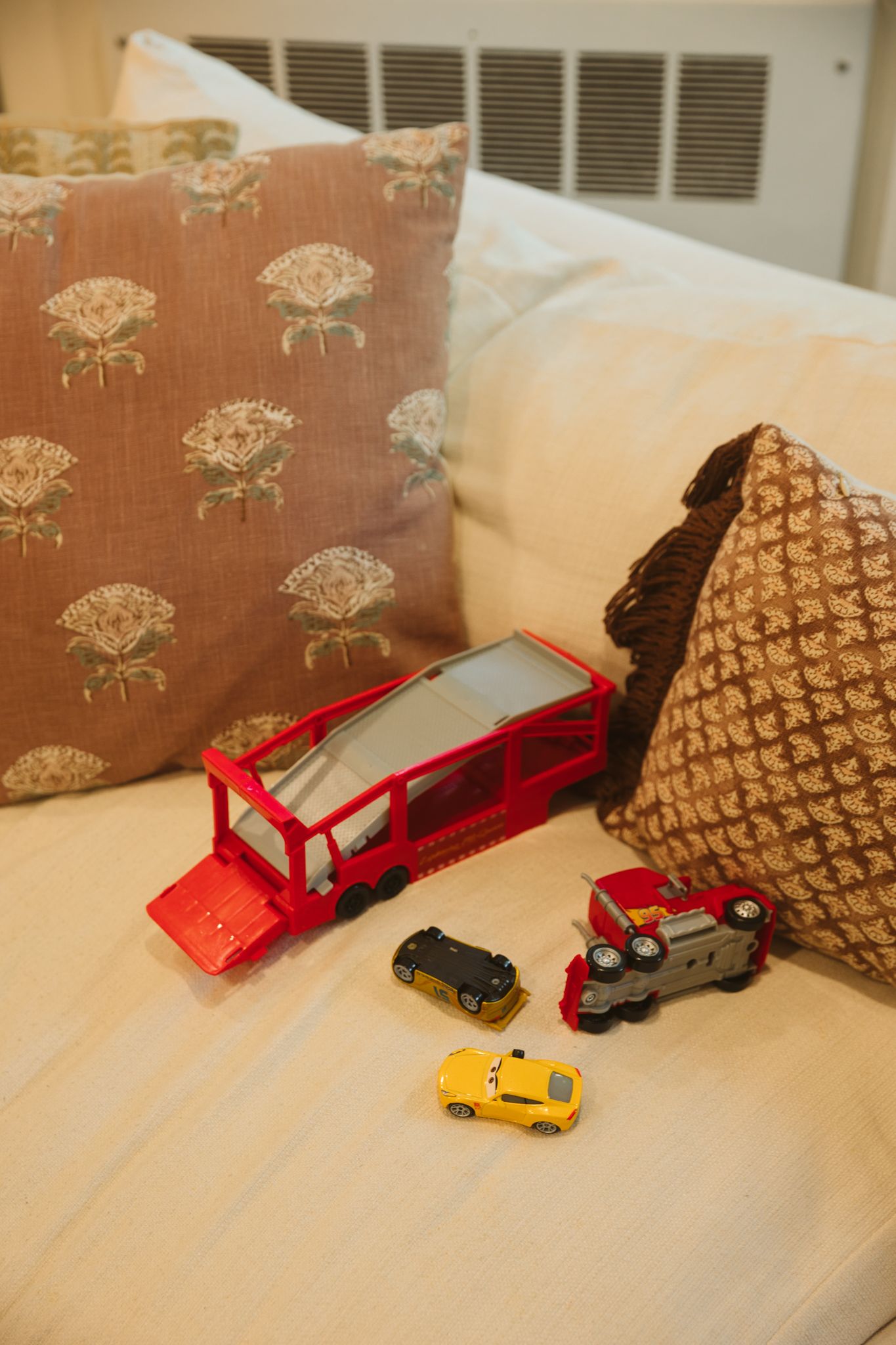 Red toy car carrier truck with small die-cast cars on a cream sofa with floral and knit decorative pillows.
