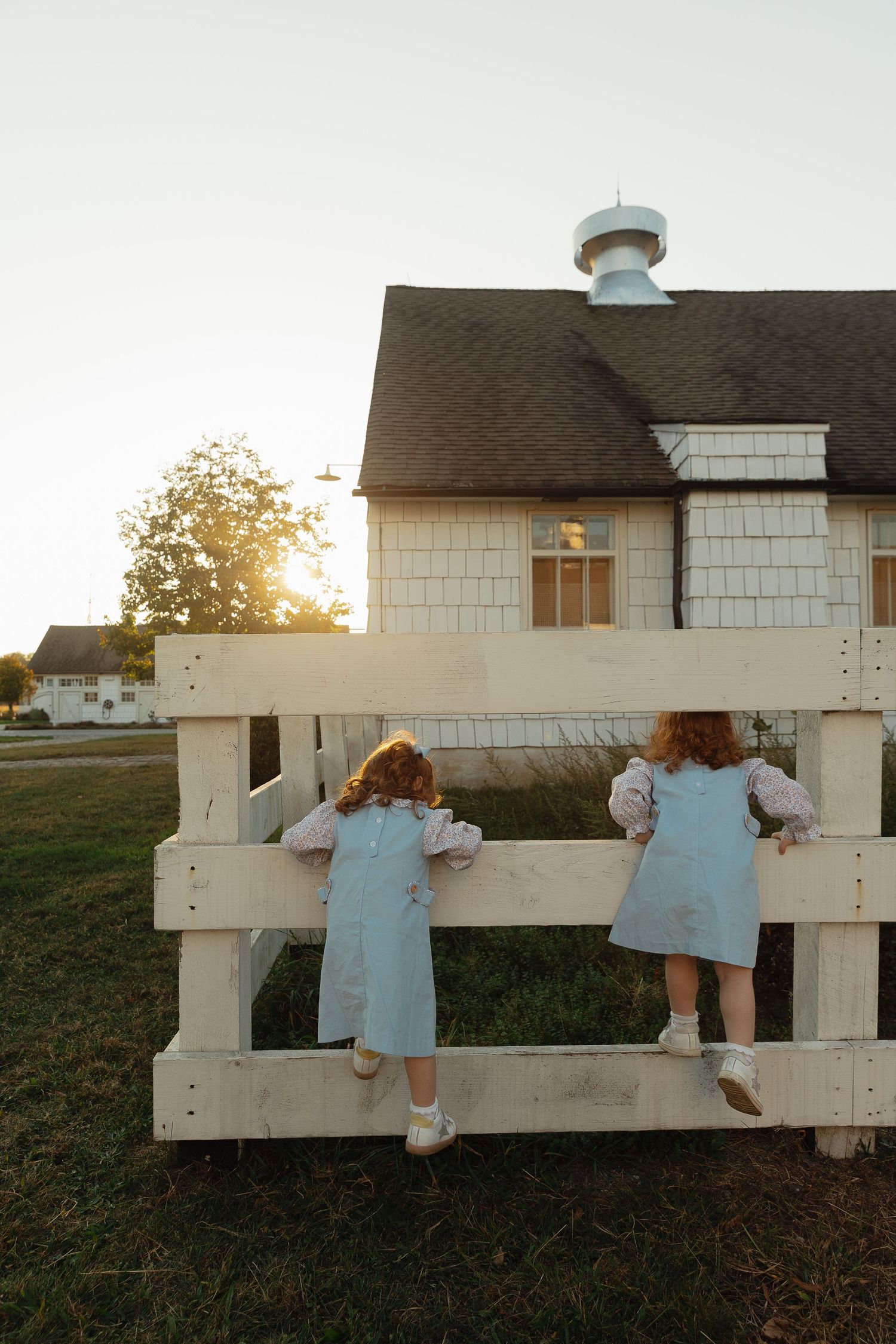 Two young redhead girls in matching blue dresses climbing a white wooden fence at golden hour near a farmhouse.