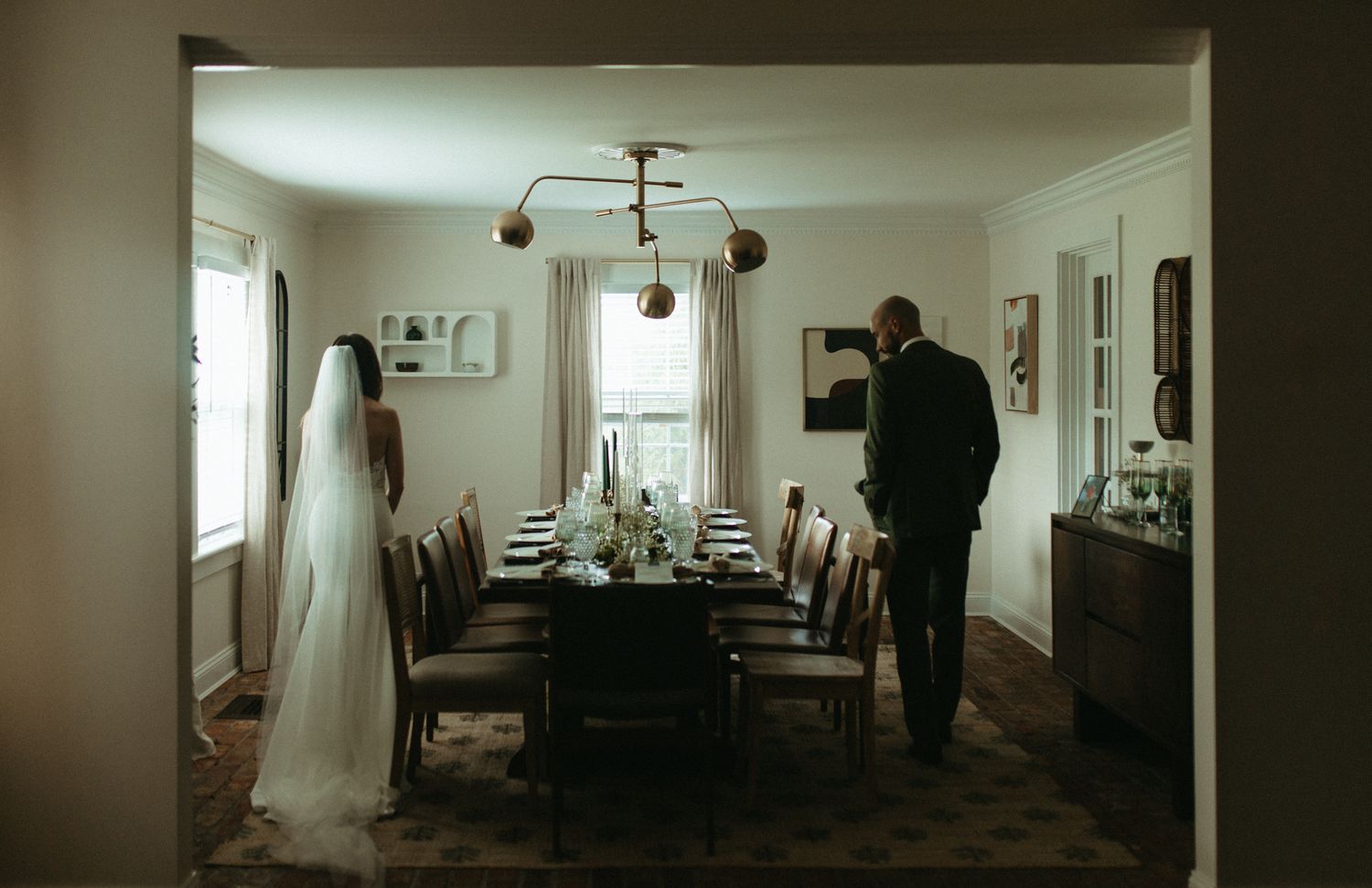 Bride and groom stand in elegant dining room with long table, floral centerpiece, and chandelier on their wedding day.