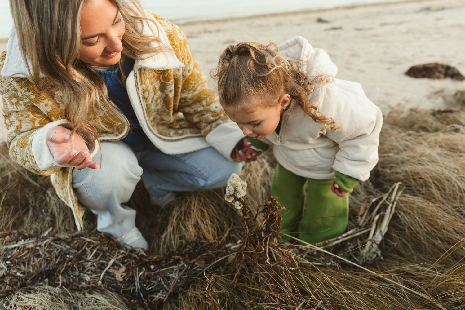 A mother and young daughter explore seaweed on a sandy beach together, both crouching down in casual outdoor clothing.