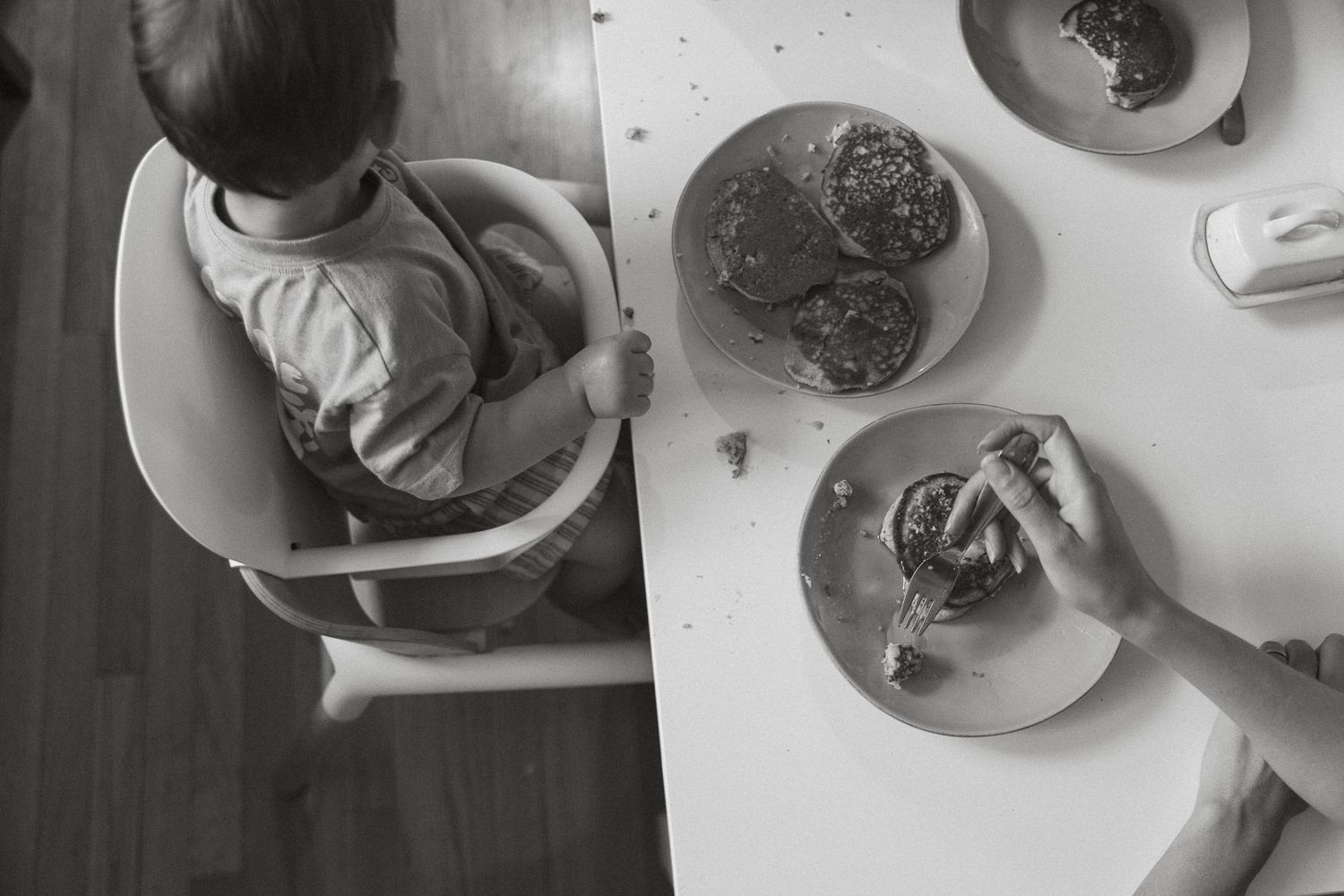 Black and white overhead photo of a toddler in a high chair at a white table with plates of food.