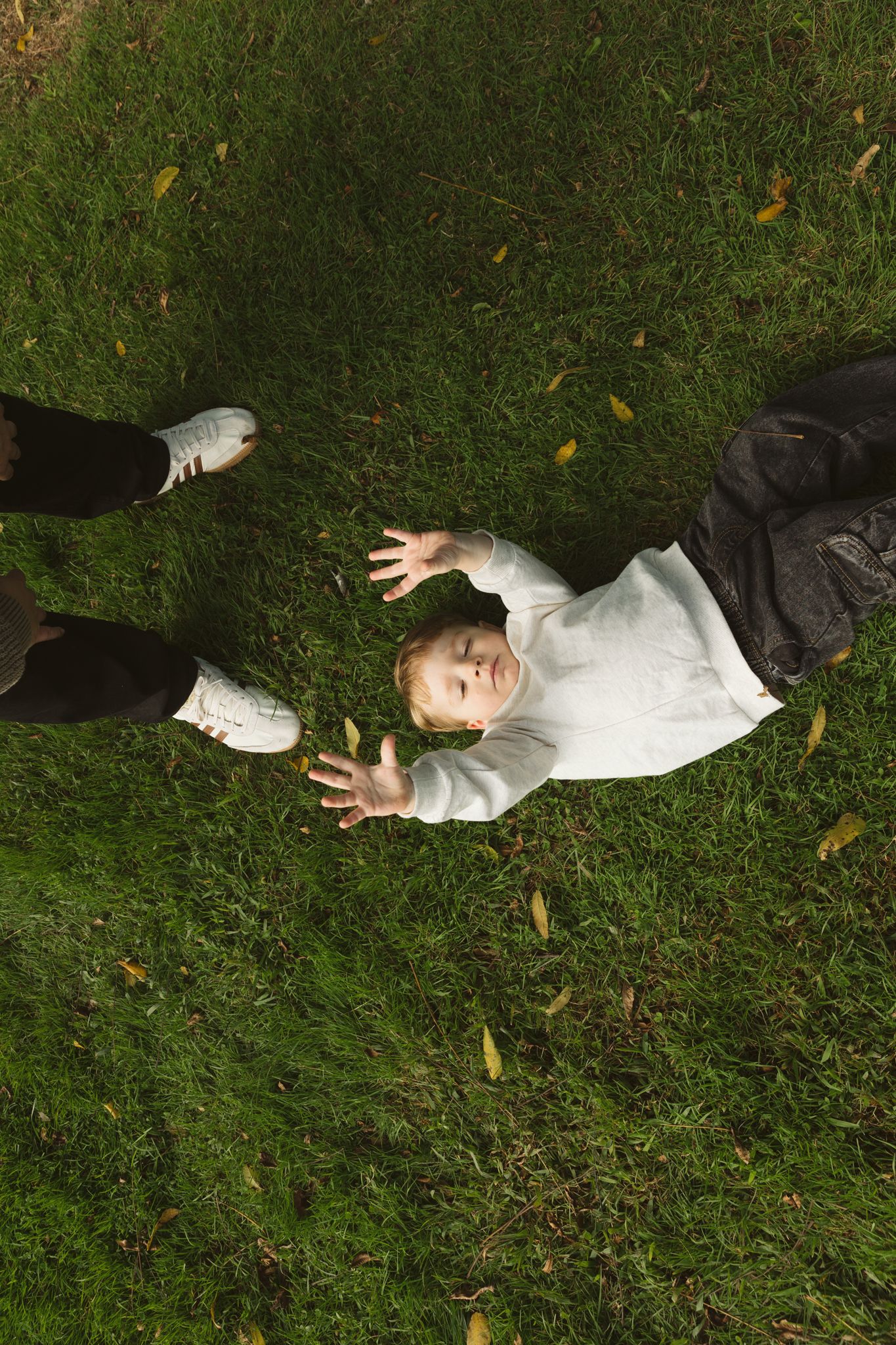 Aerial view of a baby in white sweater lying on green grass, reaching up with hands toward the camera.