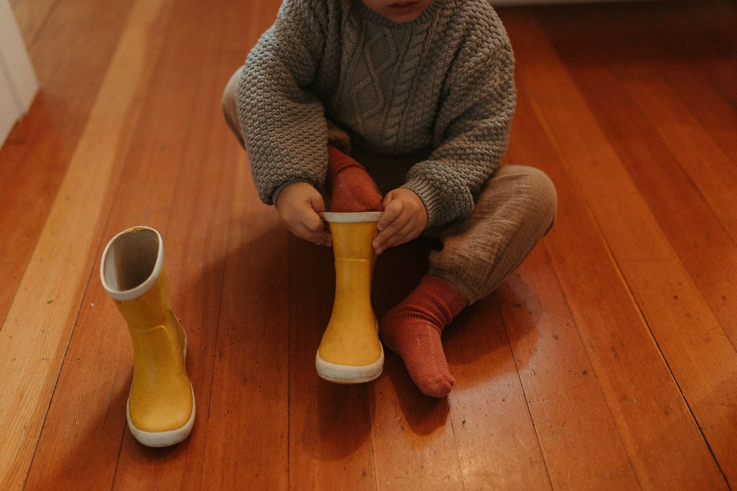Toddler in gray knit sweater sitting on hardwood floor putting on yellow rain boots.