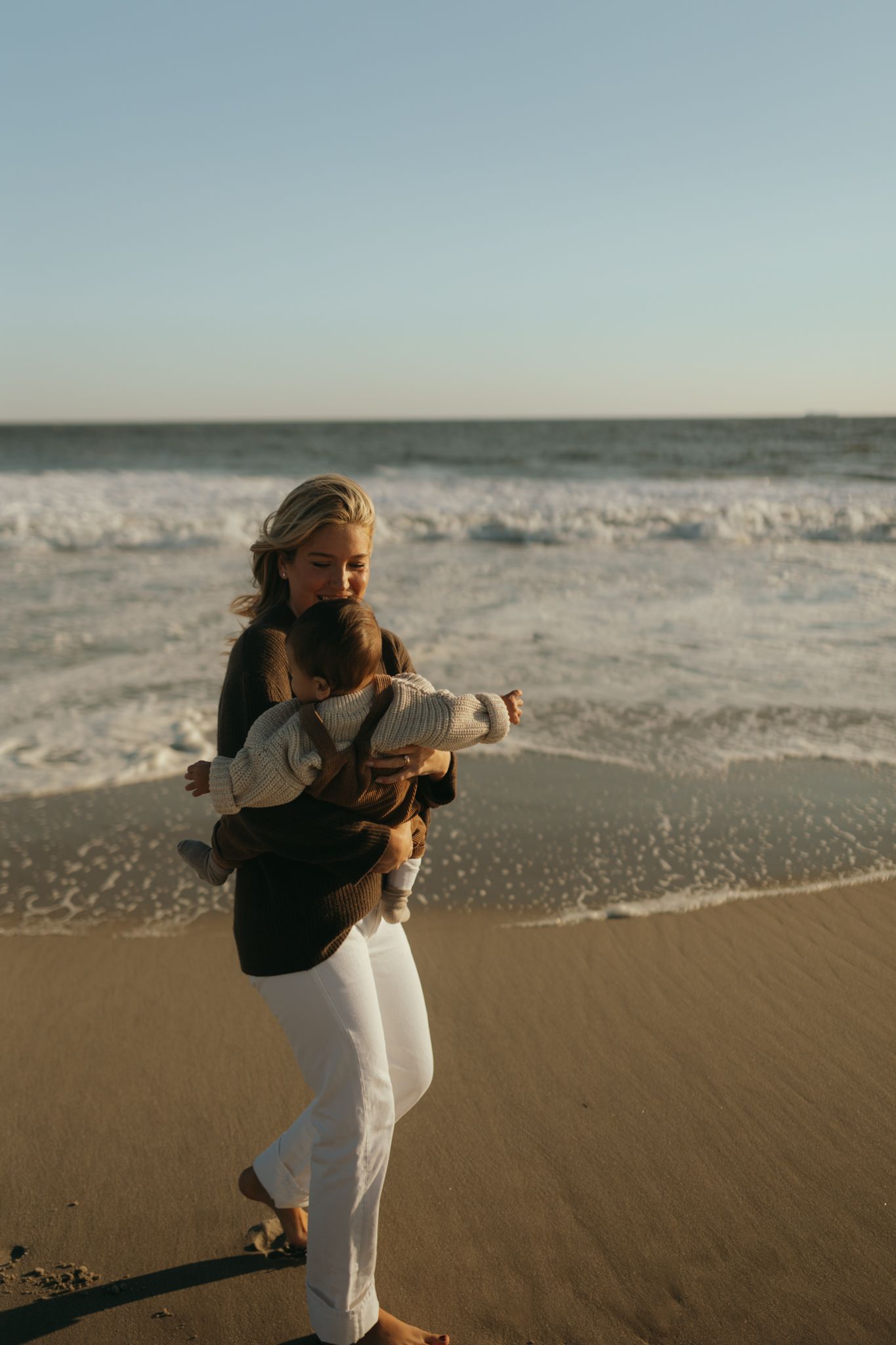 A woman in white pants stands on the beach at sunset with waves crashing behind her.
