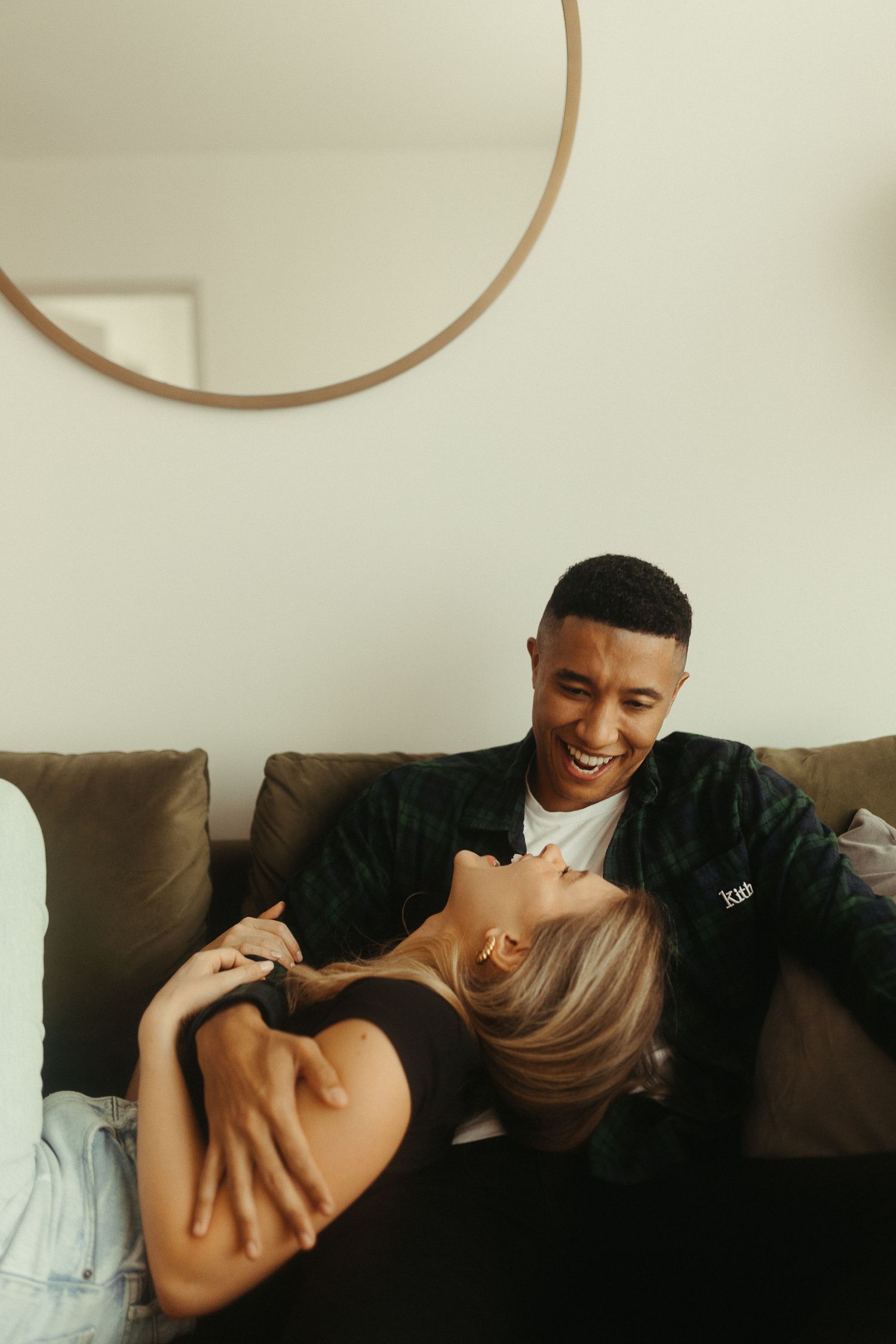 Happy interracial couple laughing together on couch beneath round mirror in modern living room.