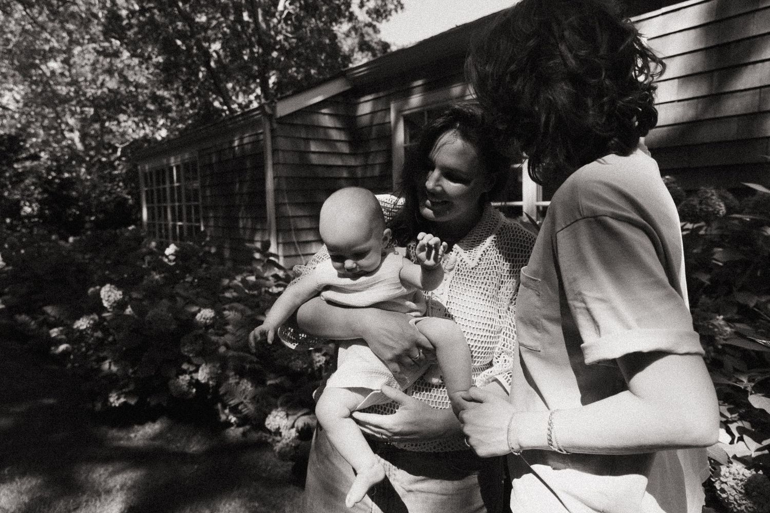 Black and white photo of two adults holding a baby outdoors near a house surrounded by lush garden foliage.