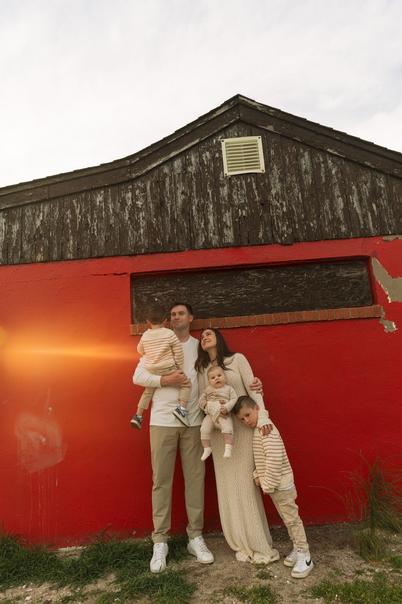 Family of four poses against a red barn wall, parents holding two young children, wearing coordinating neutral and striped outfits.