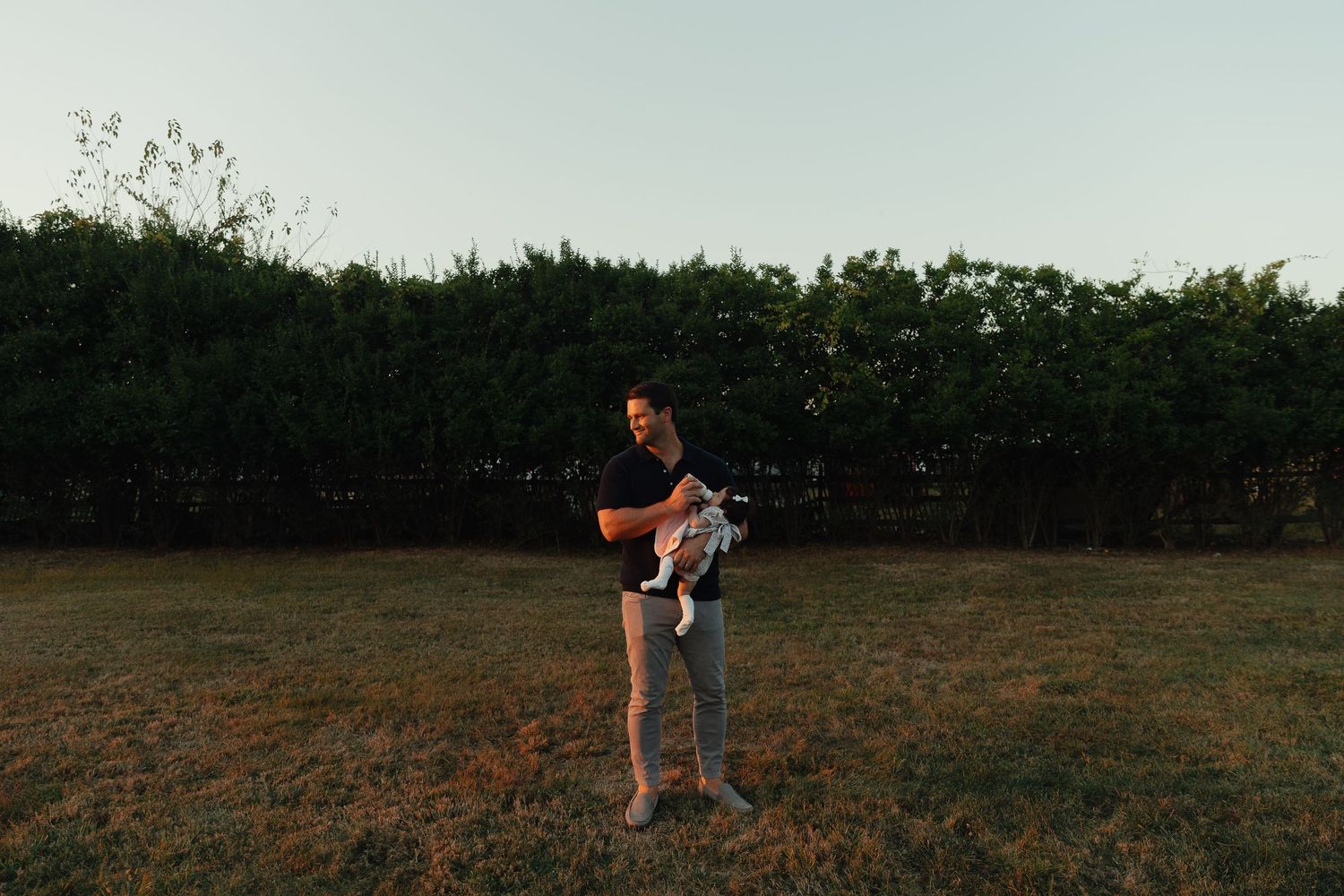 Man holding baby in arms standing on green lawn in front of tall hedge at golden hour.