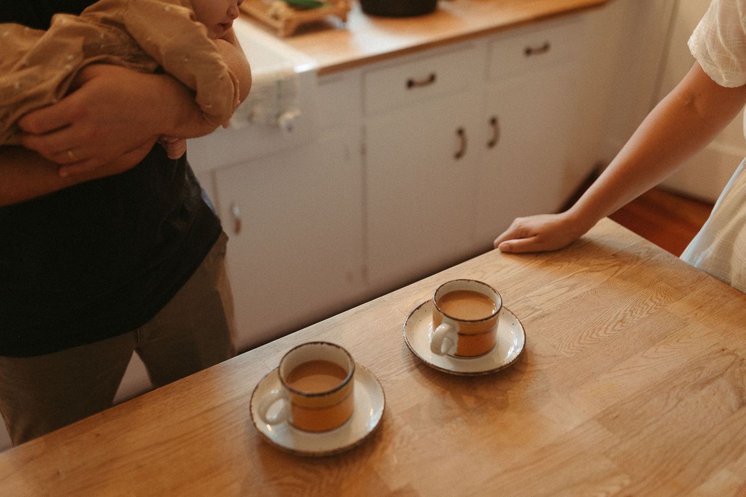 Two cups of espresso on a wooden table with adults and a baby in a cozy kitchen.