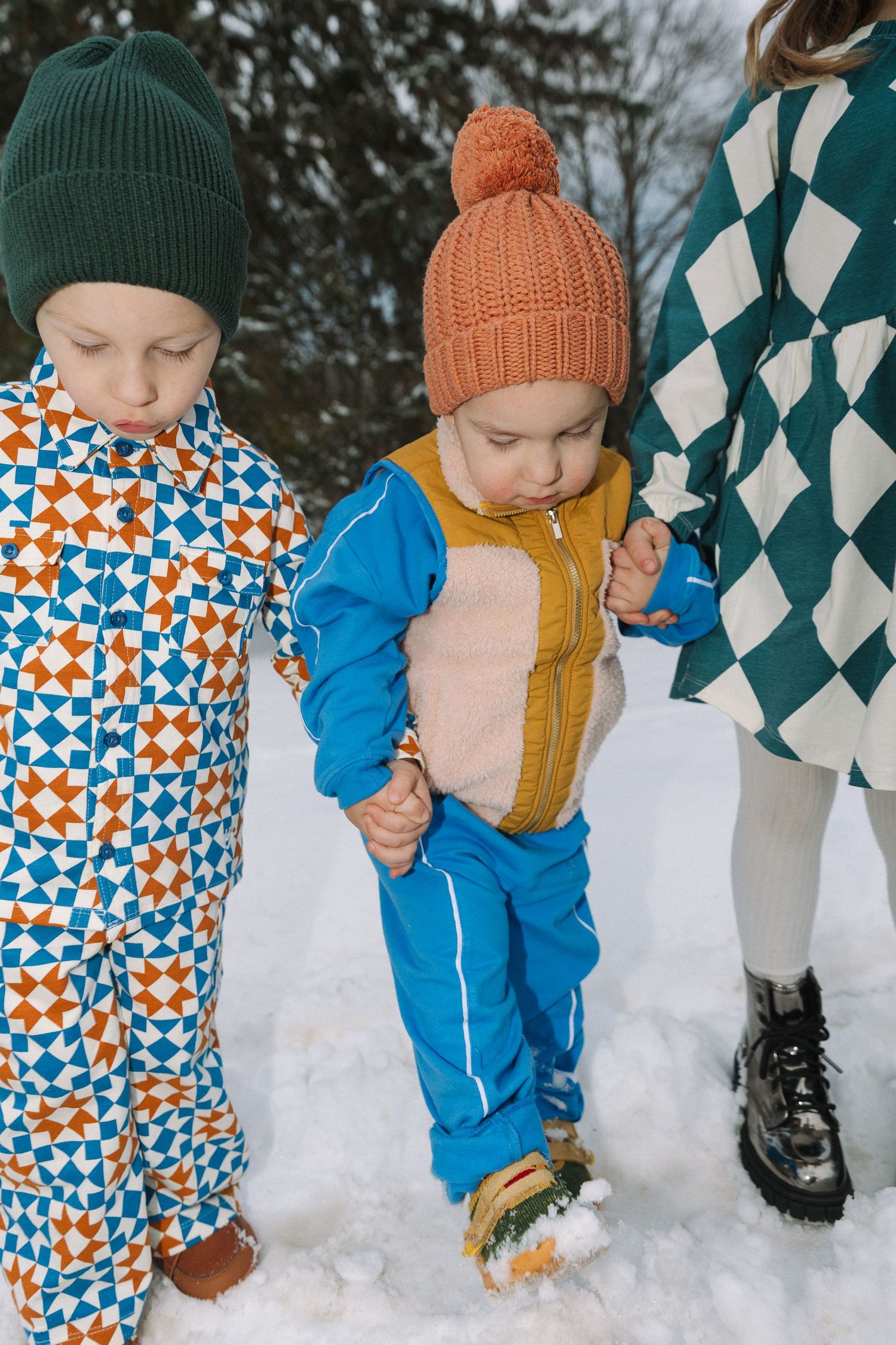Two young children in colorful winter snowsuits and hats playing in the snow, held by an adult in a green checkered coat.