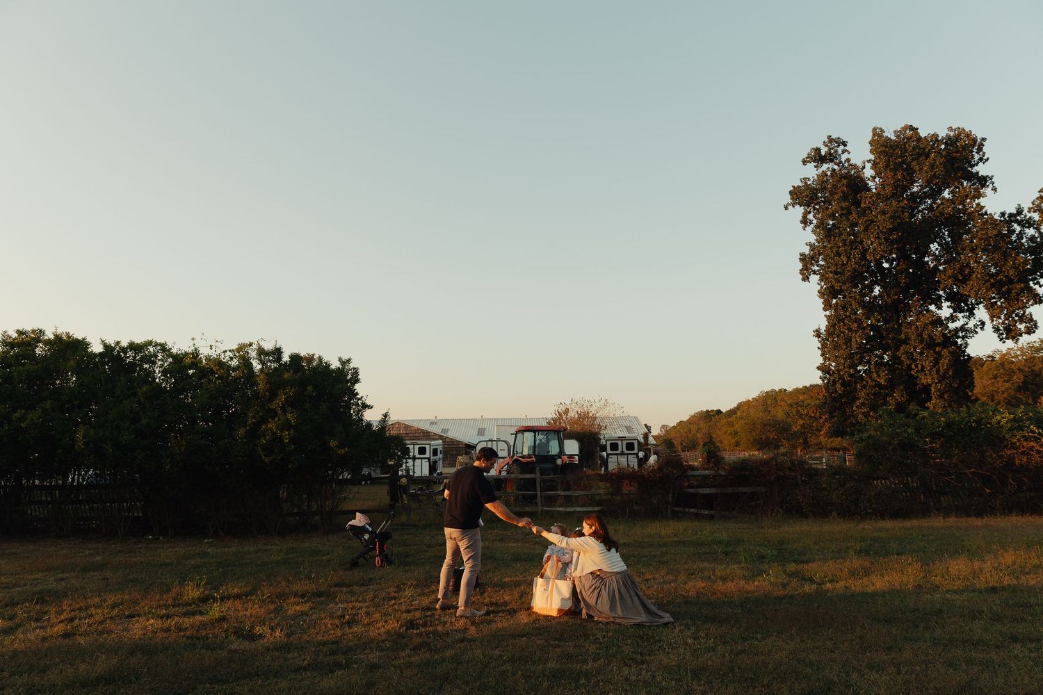 Couple holding hands at golden hour in a scenic field with trees and farm buildings in the background.