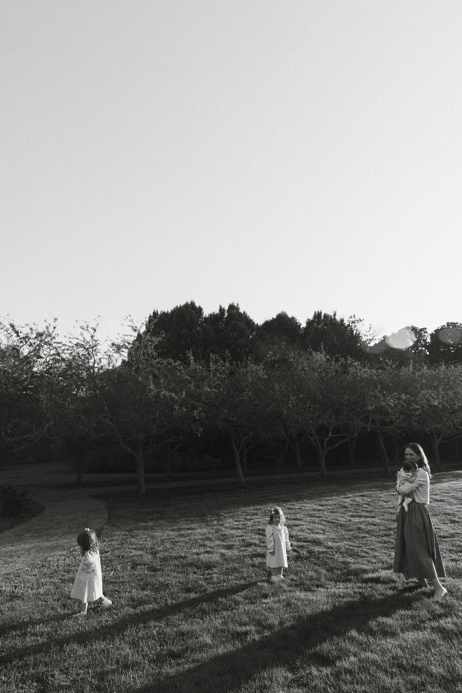 Black and white photo of a mother and two young children playing on a grassy lawn surrounded by trees.