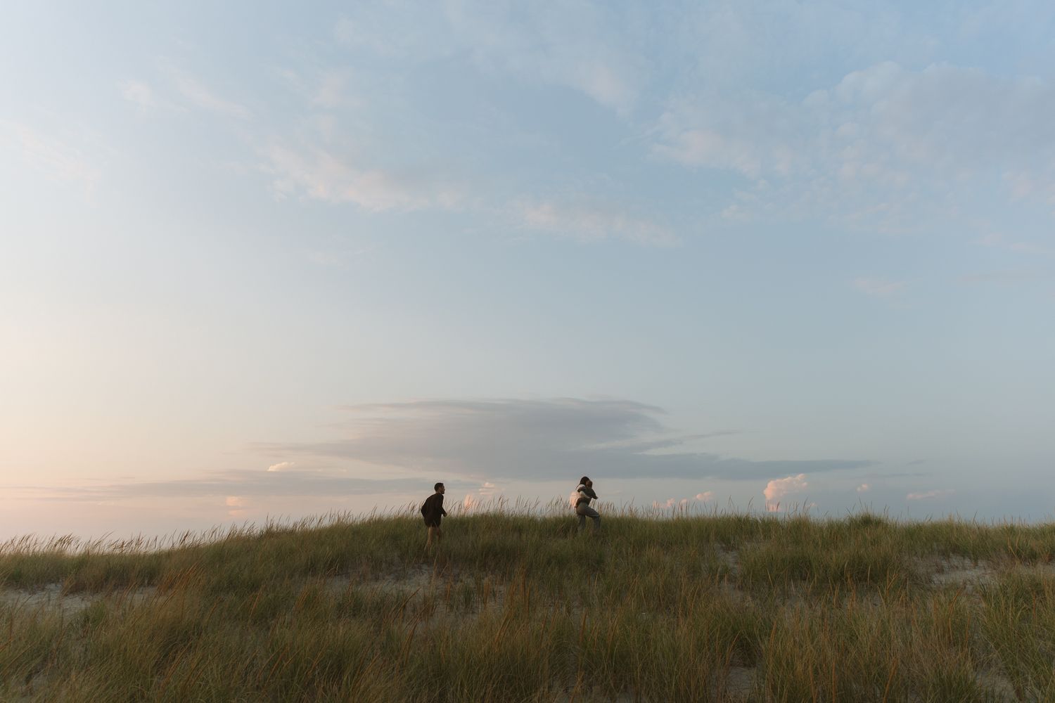 Two people walk along a grassy coastal dune at dusk under a pastel sky.