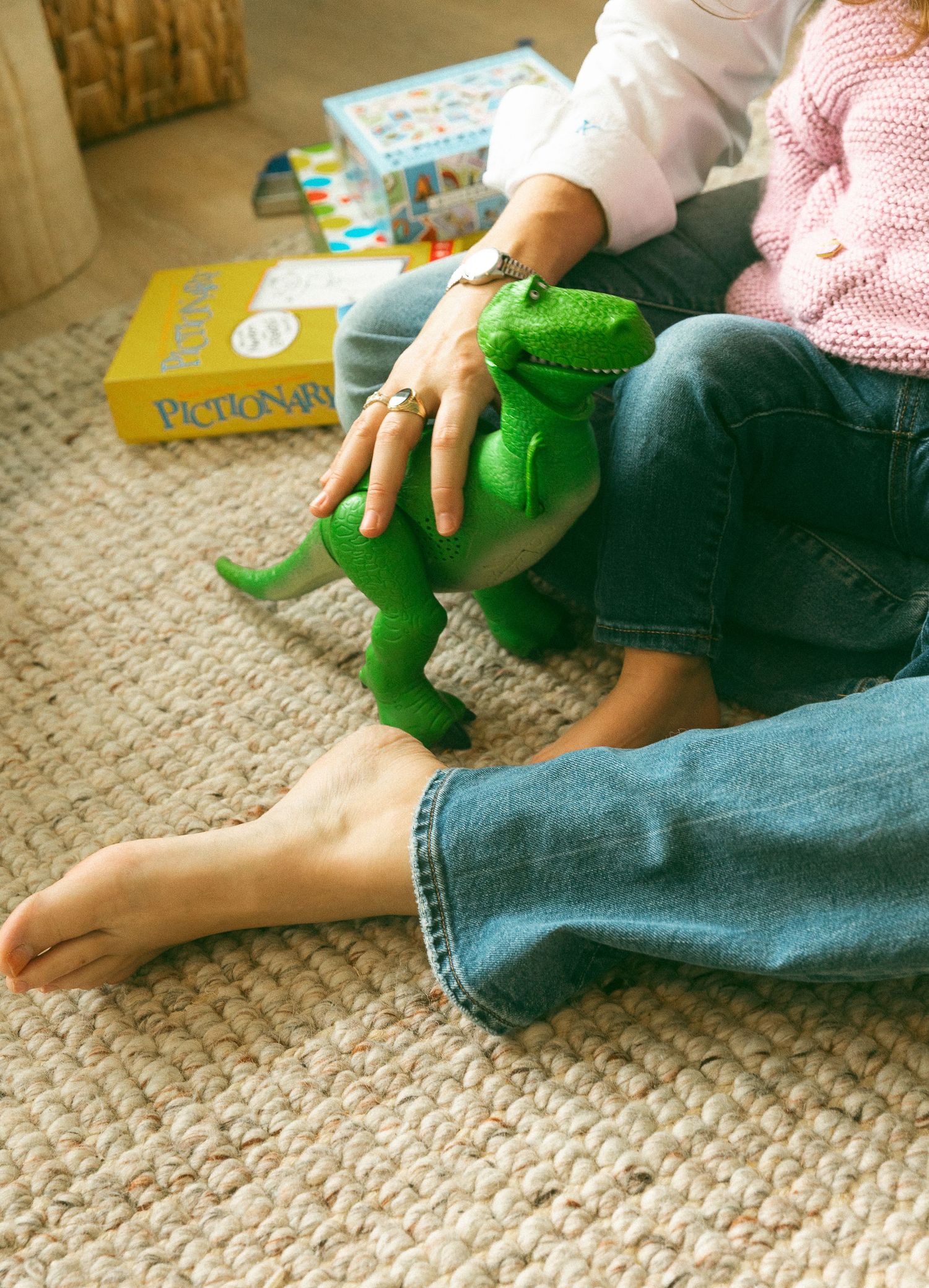 Two people sitting on a rug playing with a green T-Rex toy dinosaur, with a Pictionary board game in the background.