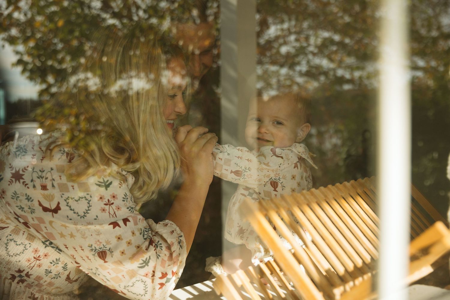 A blonde mother in a floral dress holds her smiling baby near a sunlit window with green trees outside.