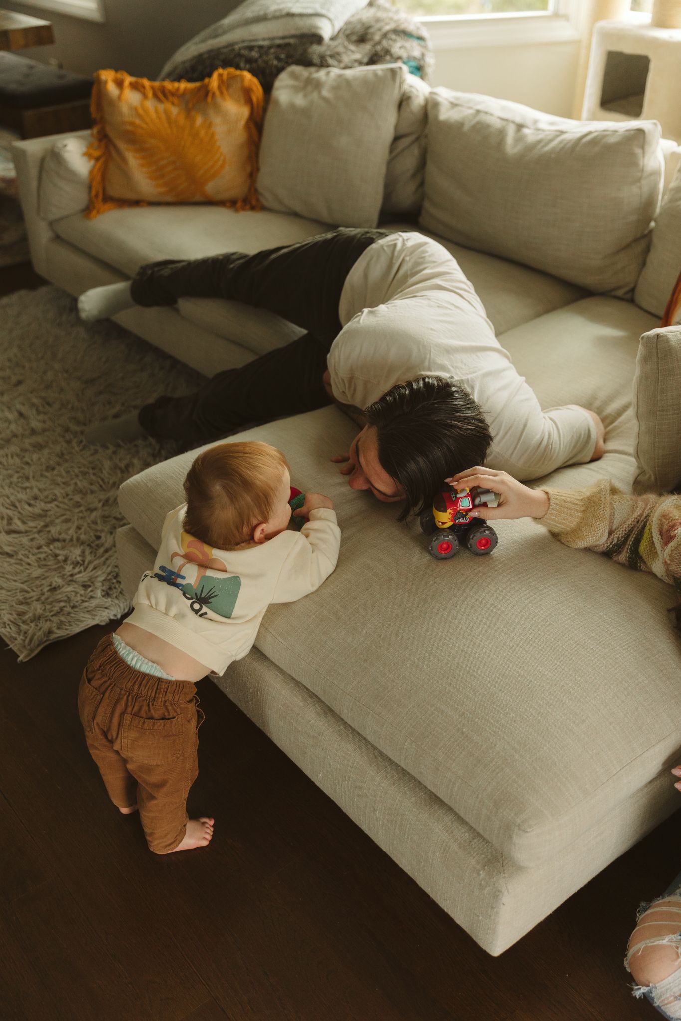 Parent and baby playing with toy trucks on a light gray sectional sofa.