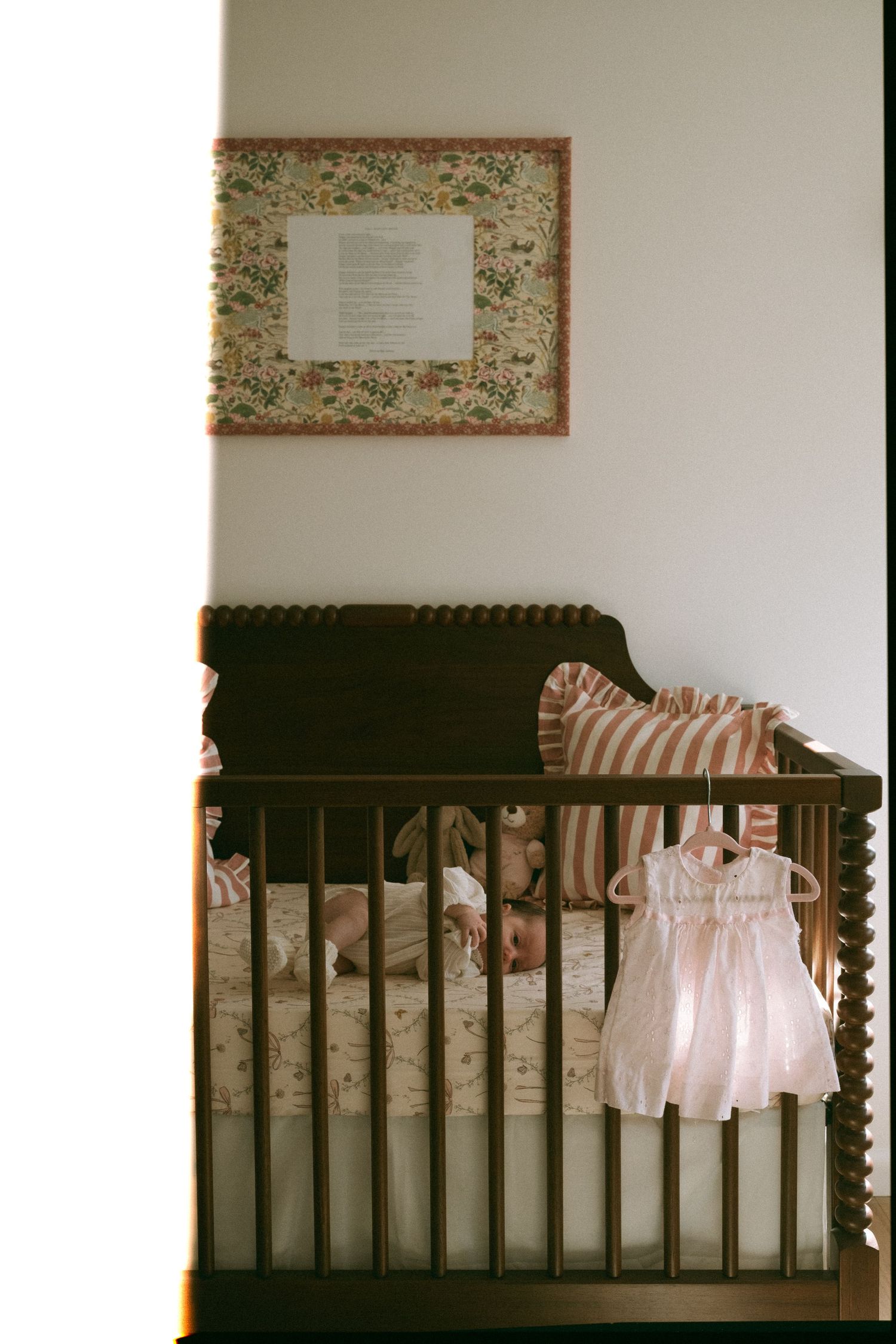 Pink baby dress hangs delicately on a dark wooden crib with floral bedding.