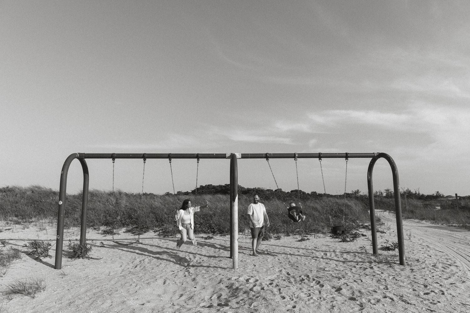 Black and white photo of swing set on sandy playground.