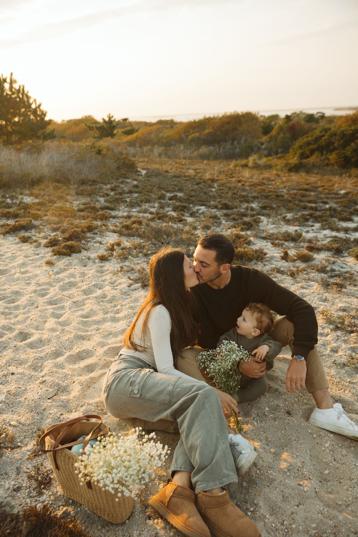 A romantic couple sitting on sandy ground with a basket of flowers at sunset.