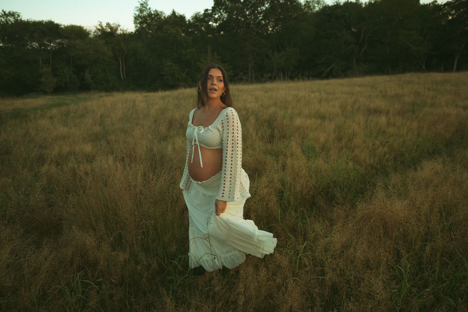 Pregnant woman in white crochet outfit stands laughing in golden sunlit meadow at sunset.