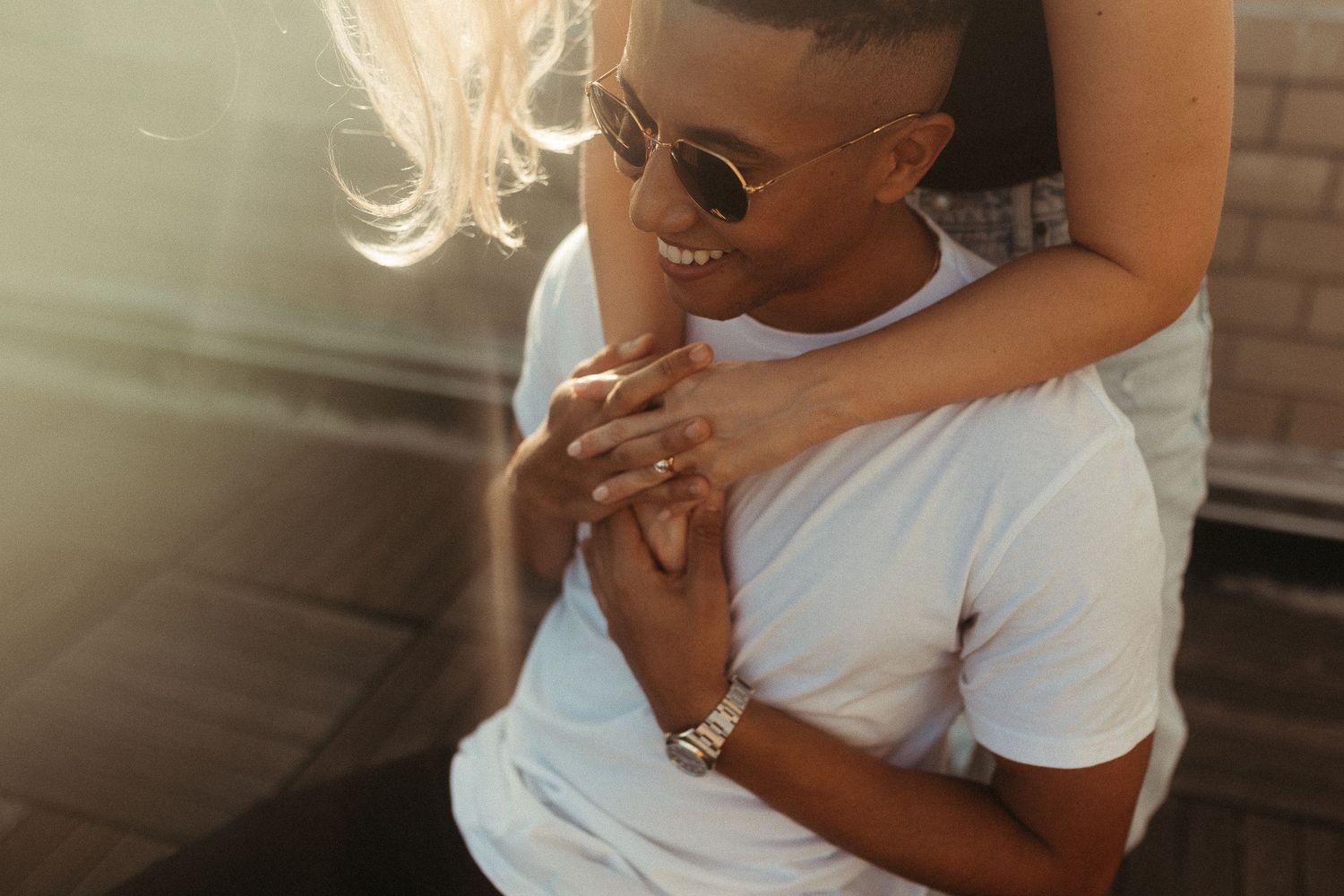 Smiling man in white t-shirt and sunglasses embraced from behind by woman in warm sunlight outdoors.