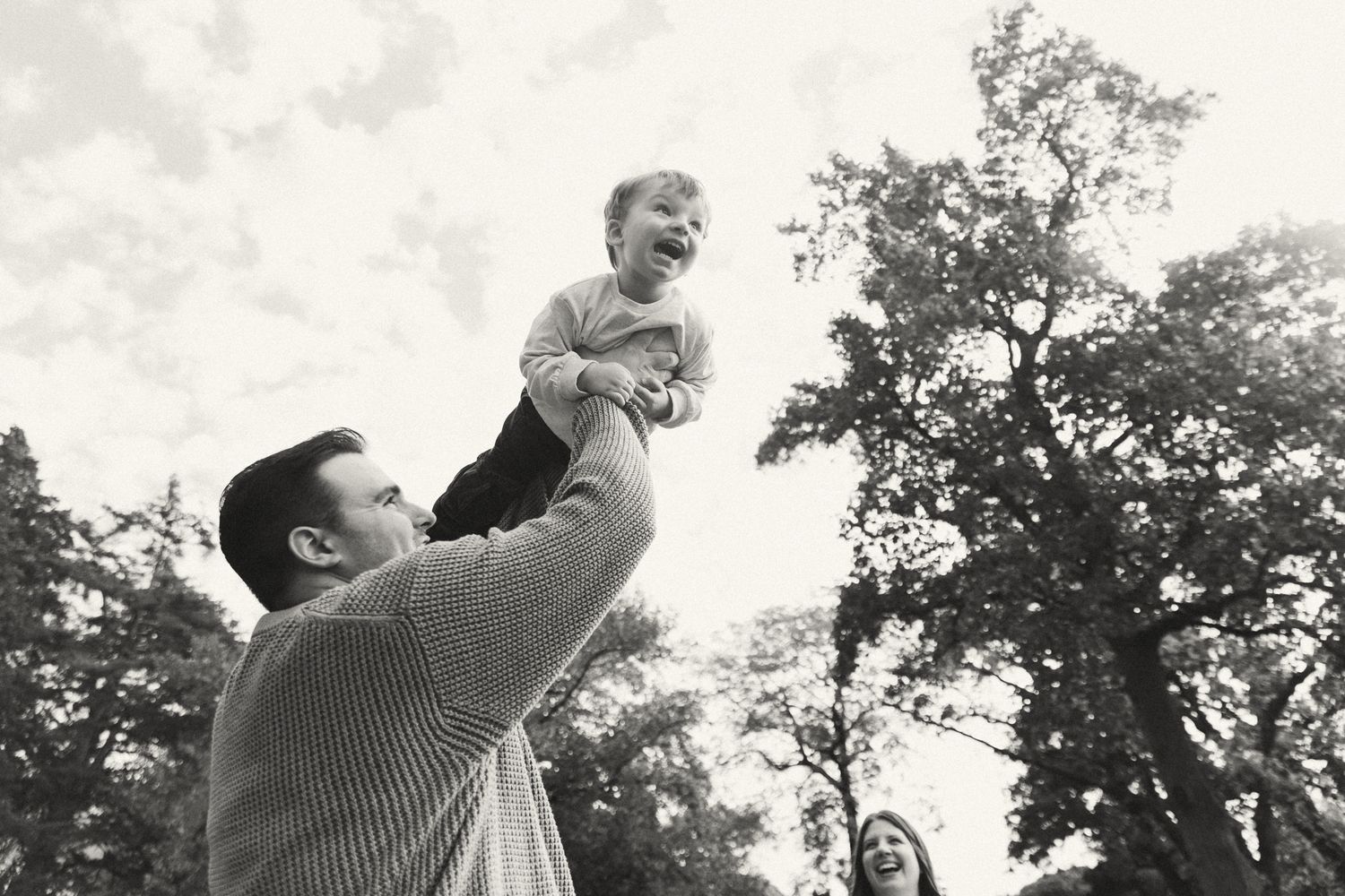 Black and white photo of a father joyfully lifting his laughing child in the air outdoors with trees in background.