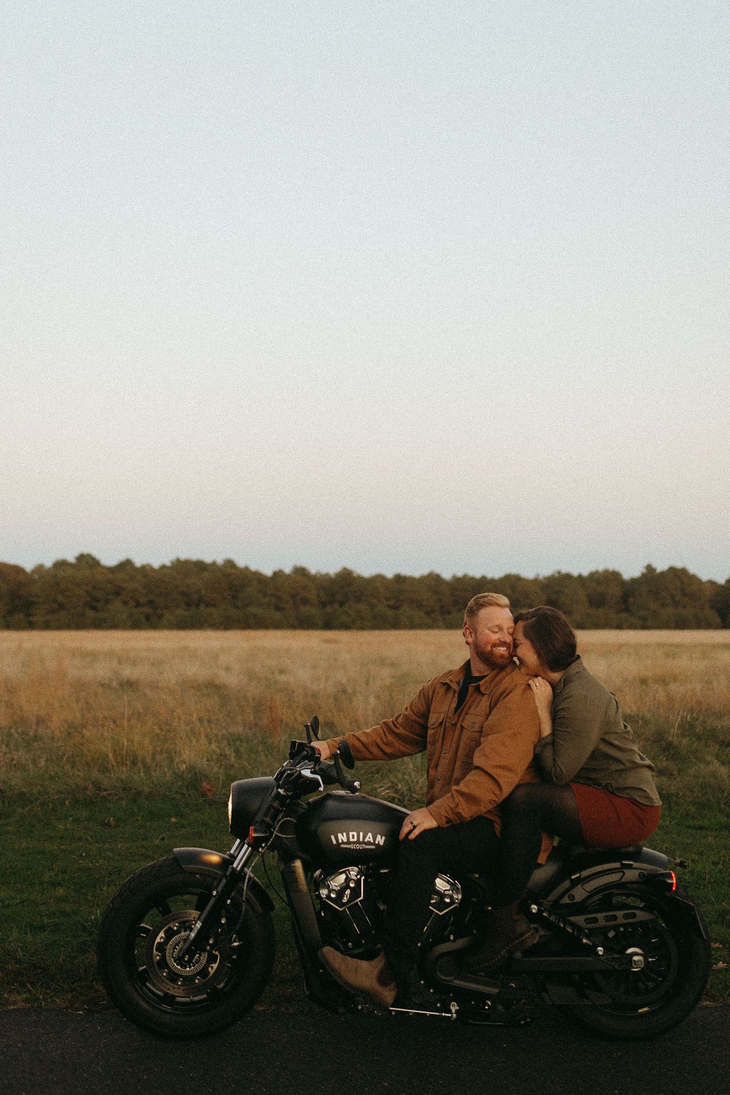 Couple sharing a romantic kiss on a black Indian motorcycle in a scenic field at golden hour.