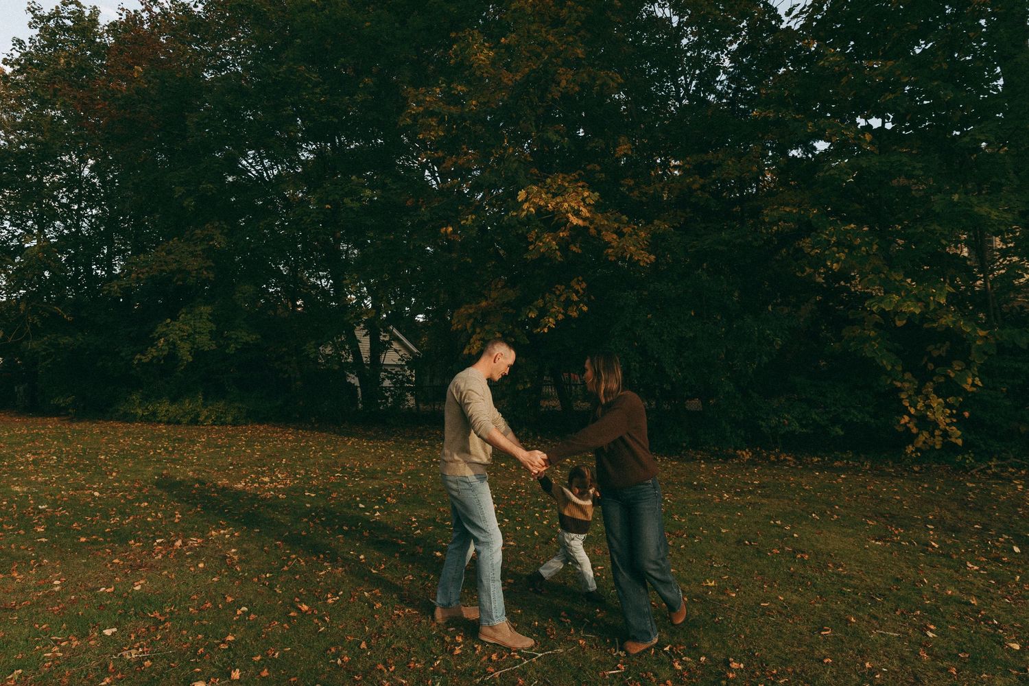 Two adults swing a toddler between them while walking through a park with autumn foliage in the background.