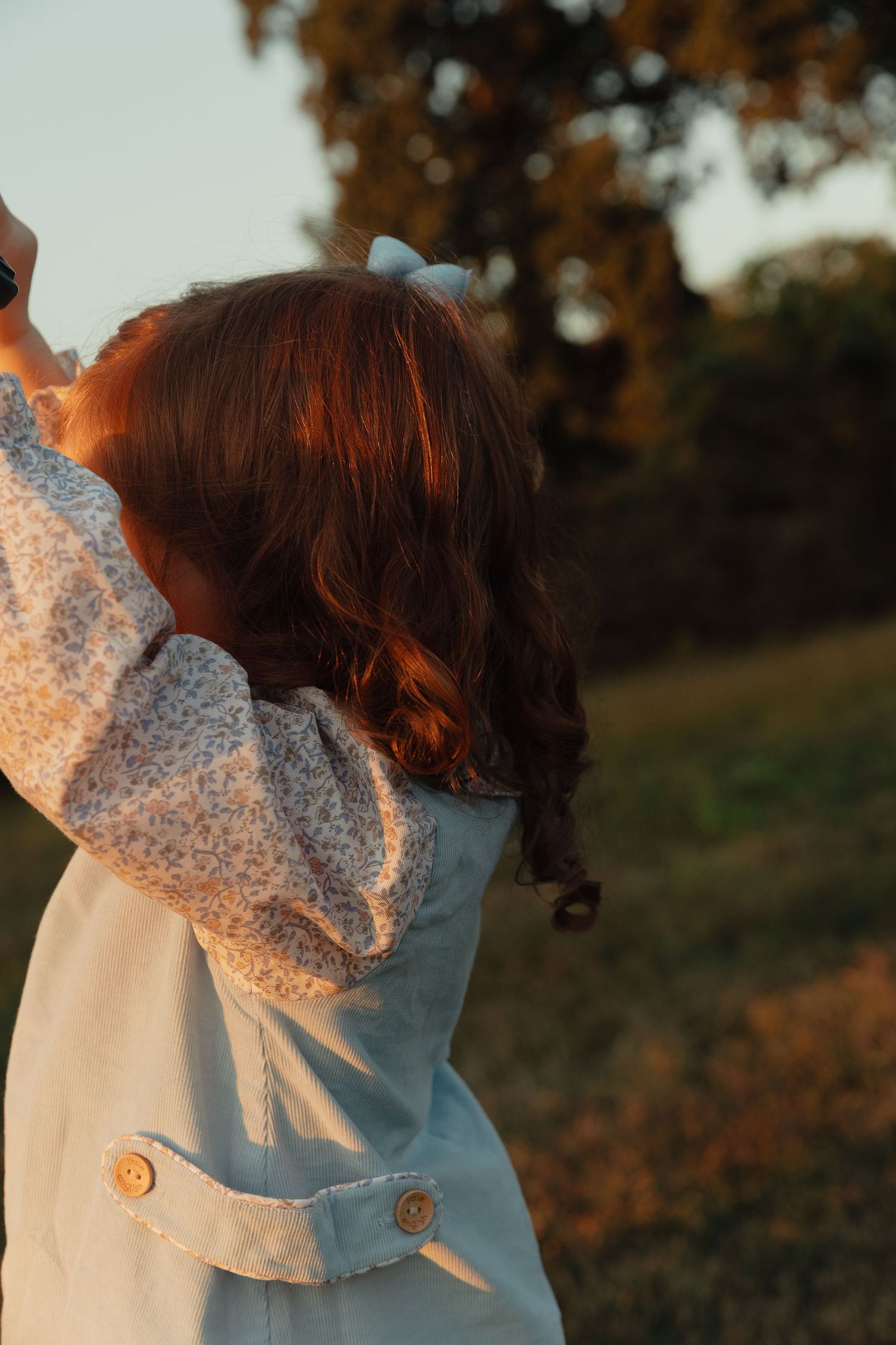 Young girl with red hair and white bow outdoors at golden hour, wearing floral blouse and light blue dress.