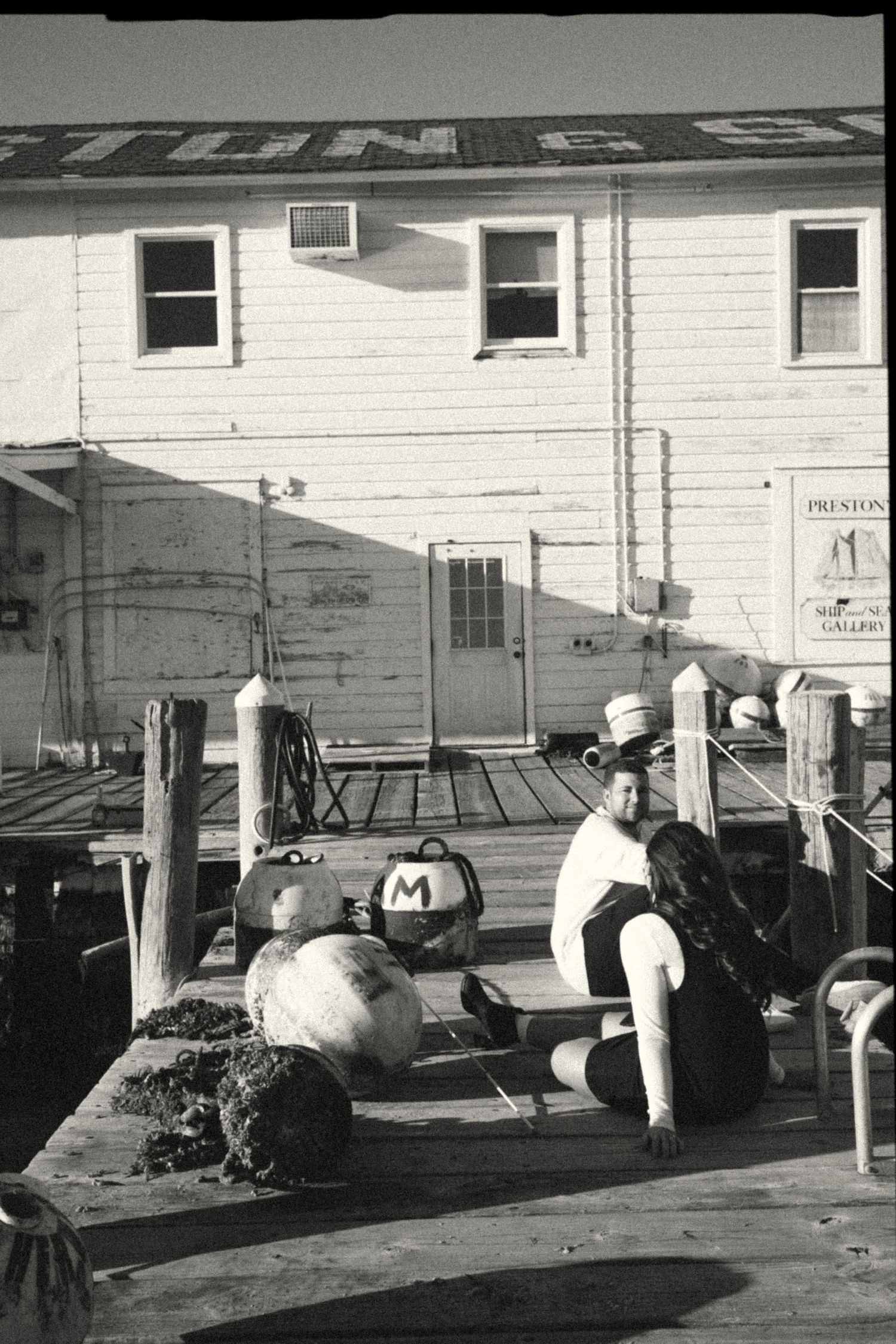 Black and white photo of a person sitting on a wooden dock near buoys, with a white building in the background.