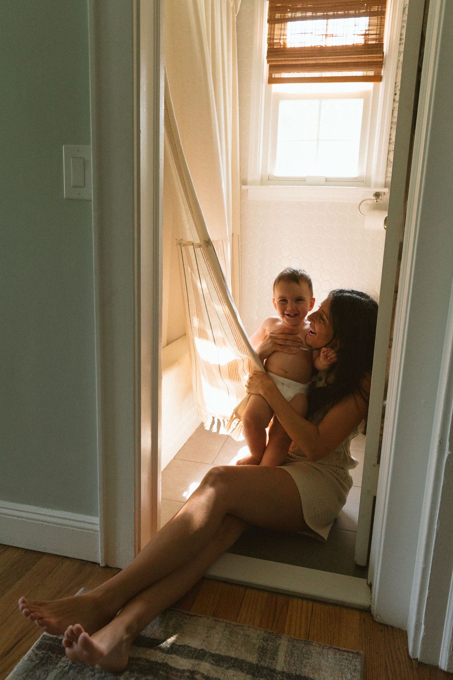 Mother sits on bathroom floor holding baby in warm window light through open doorway.