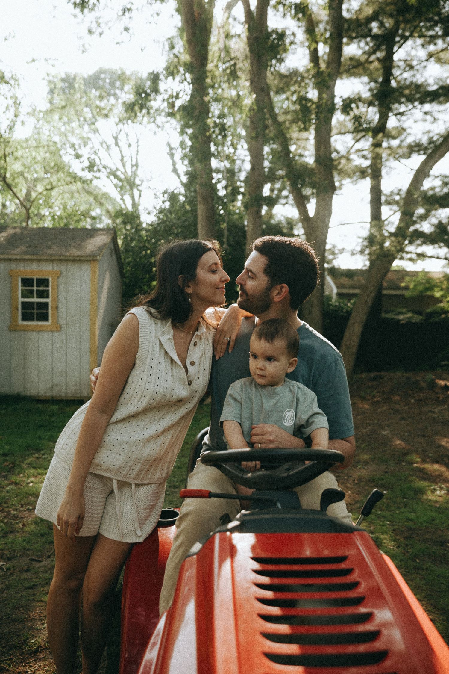 Family shares playful moment together on red riding lawn mower outdoors.
