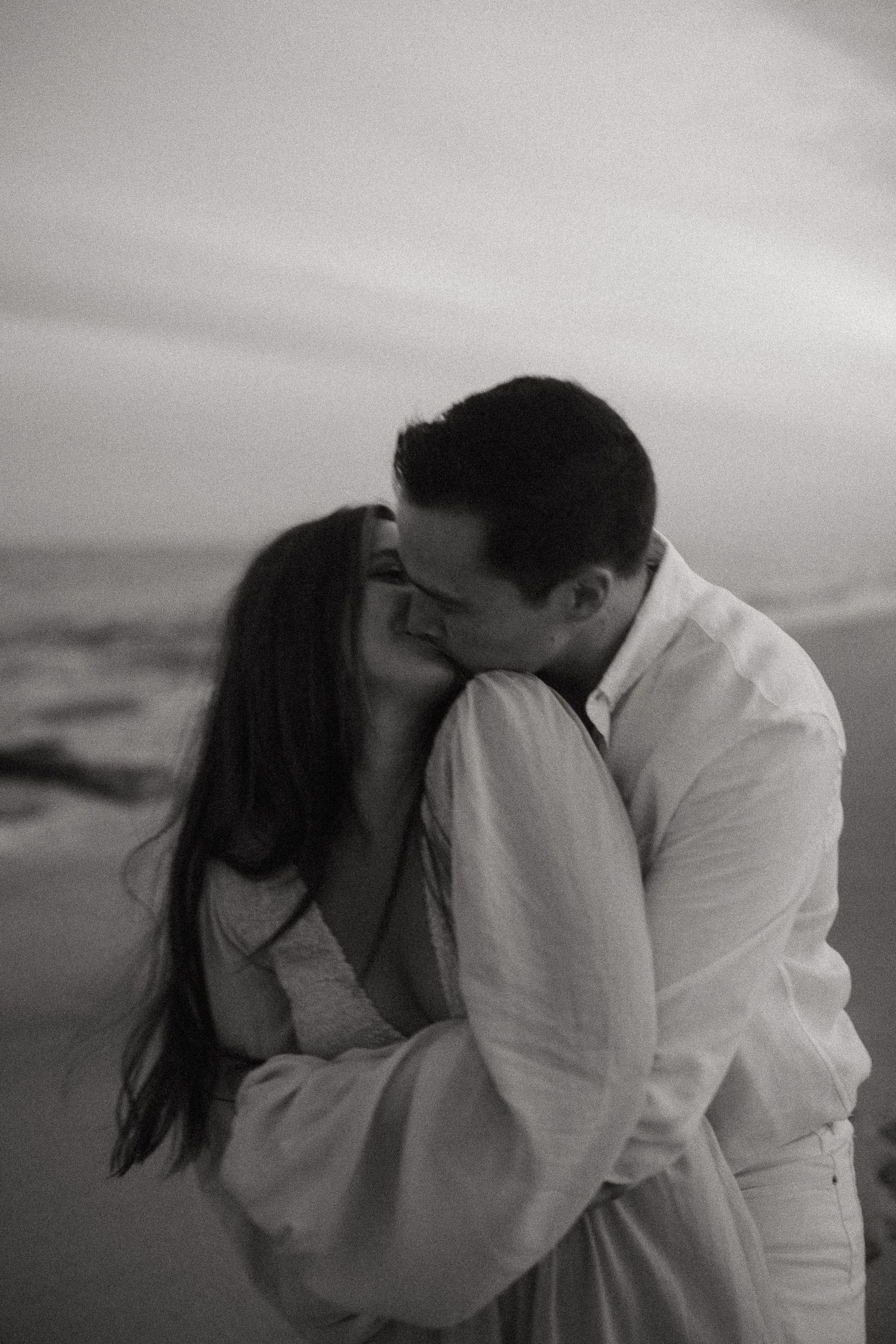 Black and white romantic photo of a couple kissing on a beach at sunset by the ocean waves.