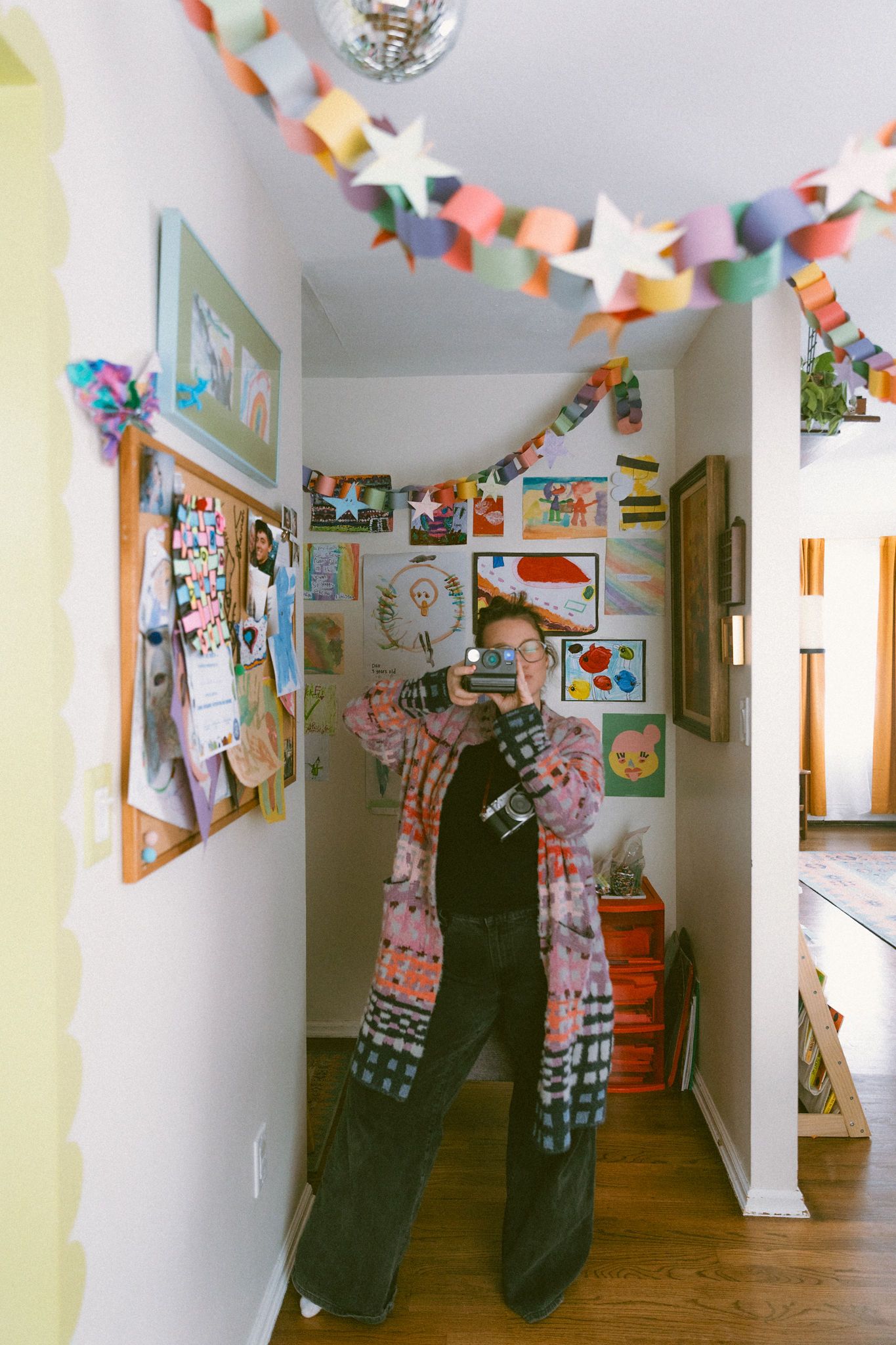 Woman photographing herself in mirror in hallway decorated with colorful childrens art and garland.