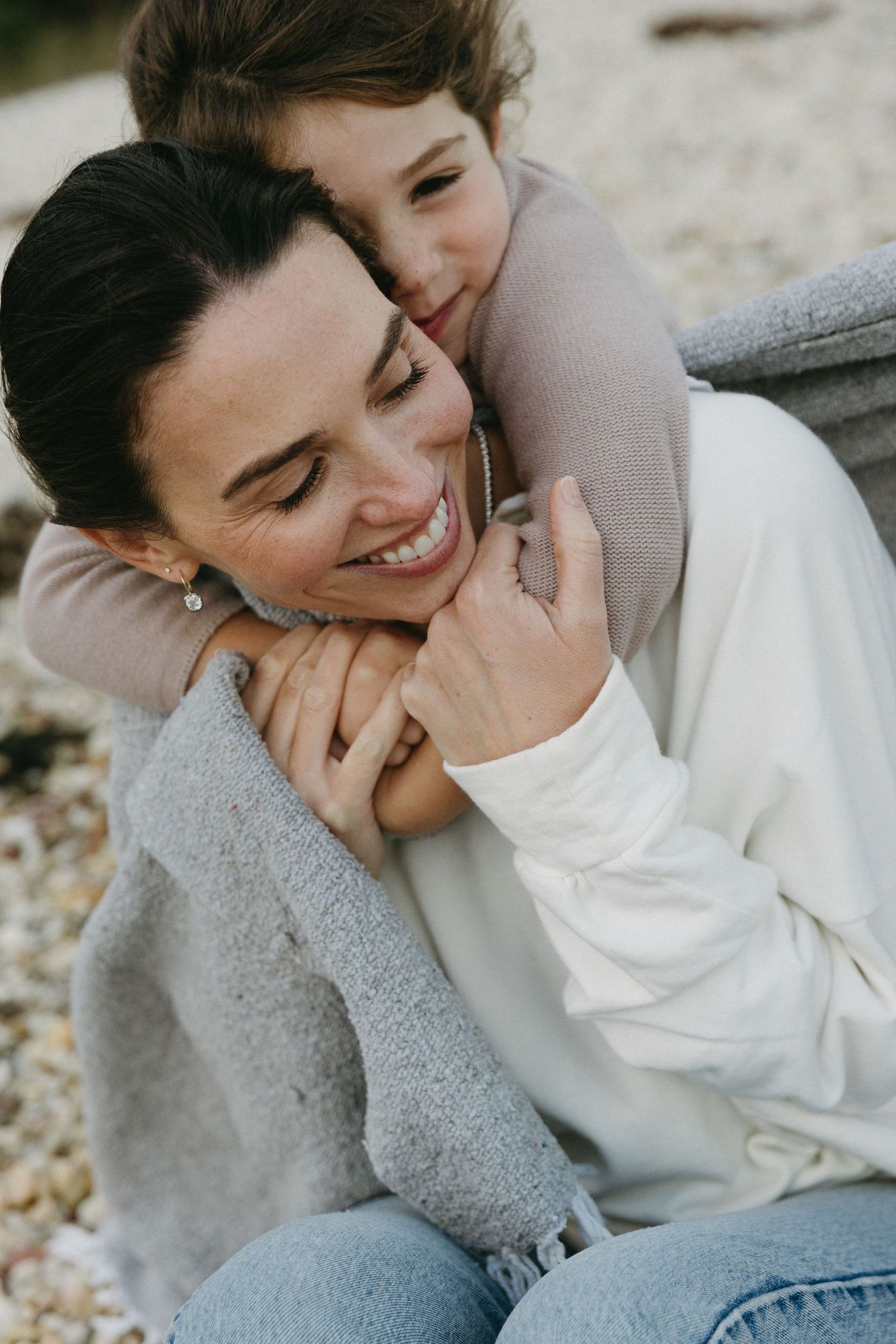 A smiling woman in a white sweater is warmly embraced from behind by a young child outdoors.